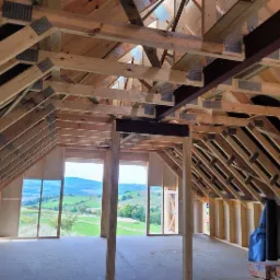 Wnętrze w stanie surowym otwartym z widokiem na pag&oacute;rkowaty krajobraz w Bielawie; drewniana konstrukcja dachu z widocznymi wiązarami i elementami stalowymi.