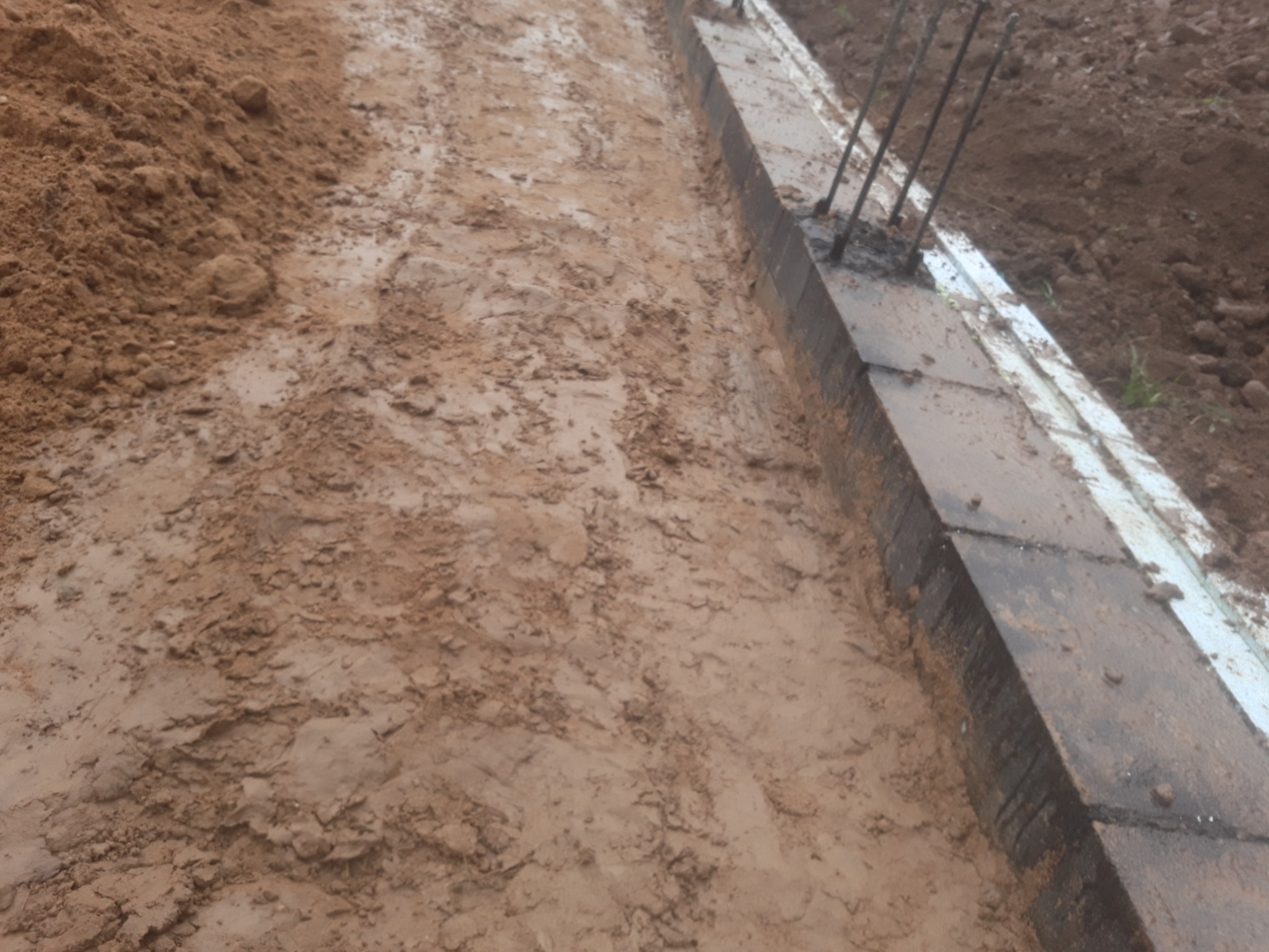 Wykop pod fundament z szalunkiem i wystającymi prętami zbrojeniowymi, ziemia pokryta błotem.