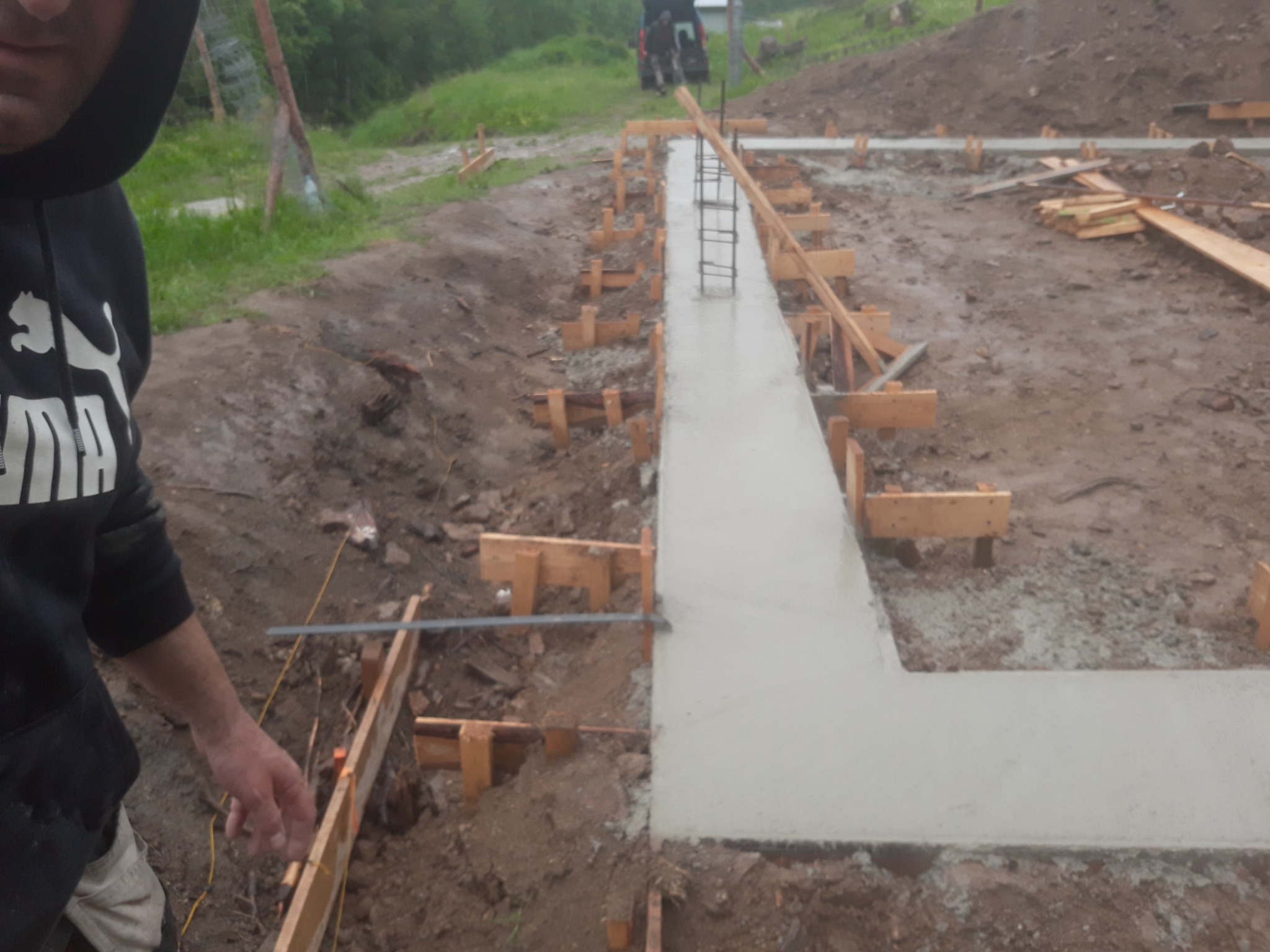 Wylany fundament zbrojony stalą, widoczne drewniane szalunki i fragment osoby w bluzie z kapturem w lewym górnym rogu.