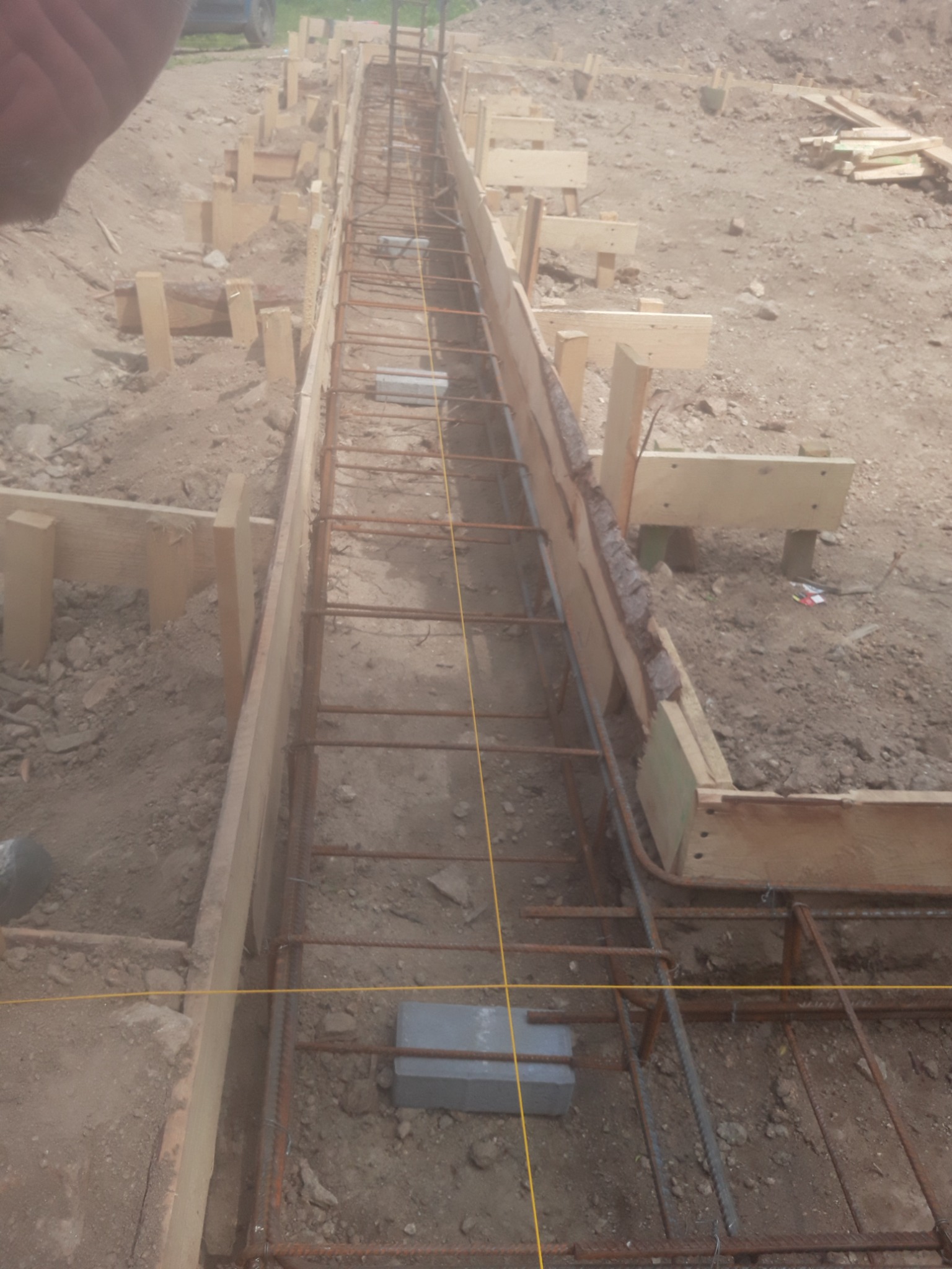 Zbrojenie w szalunku drewnianym, przygotowane do zalania betonem, widoczna linka wyznaczająca poziom, w tle ziemia i fragment samochodu.