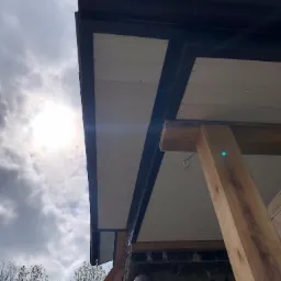 Ocieplenie elewacji z widocznymi drewnianymi elementami konstrukcyjnymi, czarnymi rynnami i białym podsufitką na tle pochmurnego nieba.