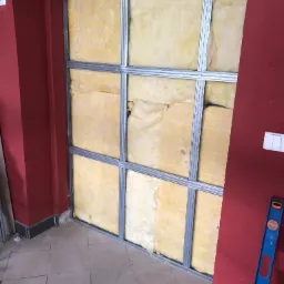 Montaż izolacji z wełny mineralnej w metalowym stelażu na tle czerwonej ściany, widoczny poziomica przy ścianie i fragment podłogi z płytek.