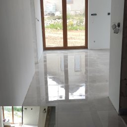 KRUGER - REMONTY KRYSTIAN KRUG - Układanie jasnych, błyszczących płytek podłogowych w nowym domu, widoczne krzyżyki dystansowe, duże okno z widokiem na ogród.