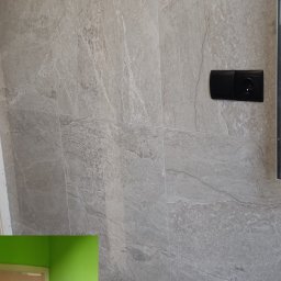 KRUGER - REMONTY KRYSTIAN KRUG - Ściana wyłożona szarymi płytkami imitującymi kamień z widocznym gniazdkiem elektrycznym. W tle fragment sąsiedniego pomieszczenia z niebieskimi płytkami i zieloną ścianą.