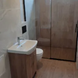 Nowoczesna łazienka z kabiną prysznicową typu walk-in z czarną ramą, umywalką na szafce z drewnopodobnym wykończeniem i toaletą podwieszaną, wykończona płytkami imitującymi drewno na podłodze...