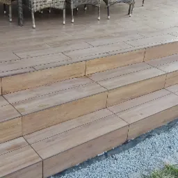 Schody zewnętrzne wyłożone płytkami imitującymi drewno, z widoczną hydroizolacją przy fundamencie i żwirowym podłożem.