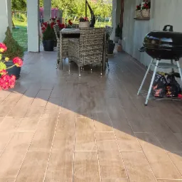 Zadaszony taras z brązowymi płytkami imitującymi drewno, na którym stoi rattanowy zestaw mebli ogrodowych i grill węglowy, widok na zielony ogród i drzewa w tle.