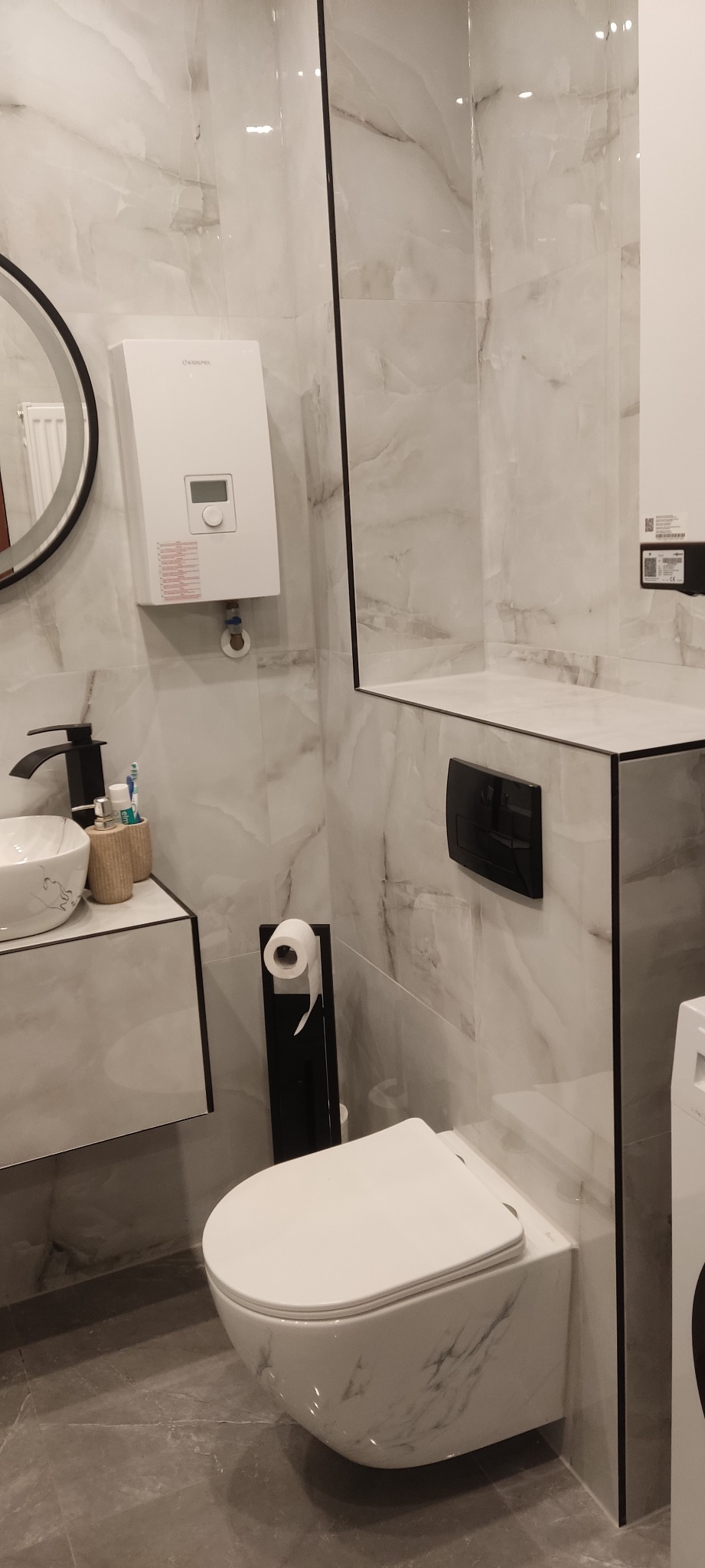 Nowoczesna łazienka z marmurowymi płytkami, podwieszaną miską WC, czarną armaturą i wiszącym podgrzewaczem wody Kospel.