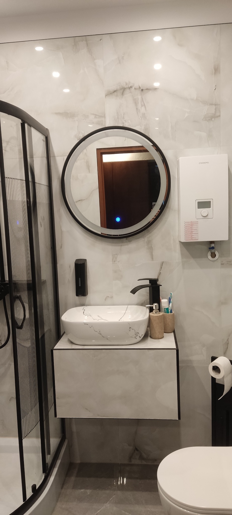 Nowoczesna łazienka z marmurowymi płytkami, okrągłym lustrem z podświetleniem LED, umywalką nablatową z czarną armaturą i kabiną prysznicową z czarnymi profilami.