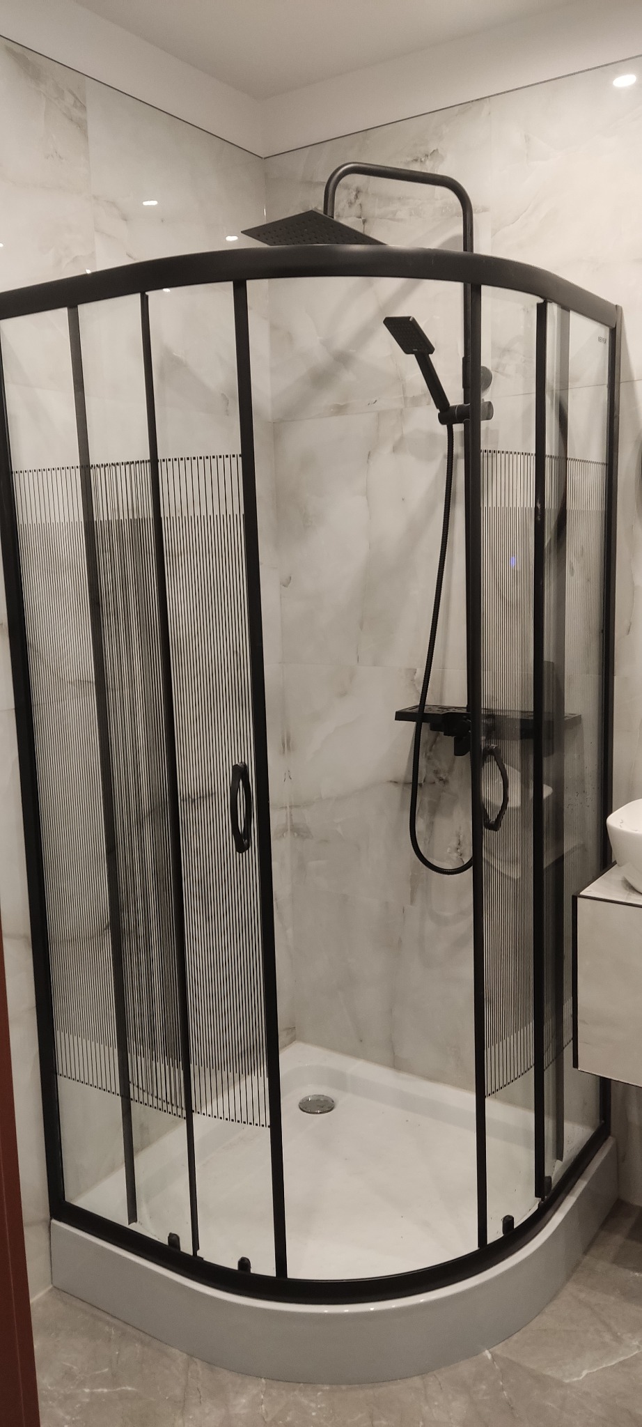 Nowoczesna kabina prysznicowa narożna z czarnymi profilami i wzorem pionowych linii na szkle, zamontowana na tle marmurowych płytek ściennych, z widocznym zestawem prysznicowym w czarnym kolorze.