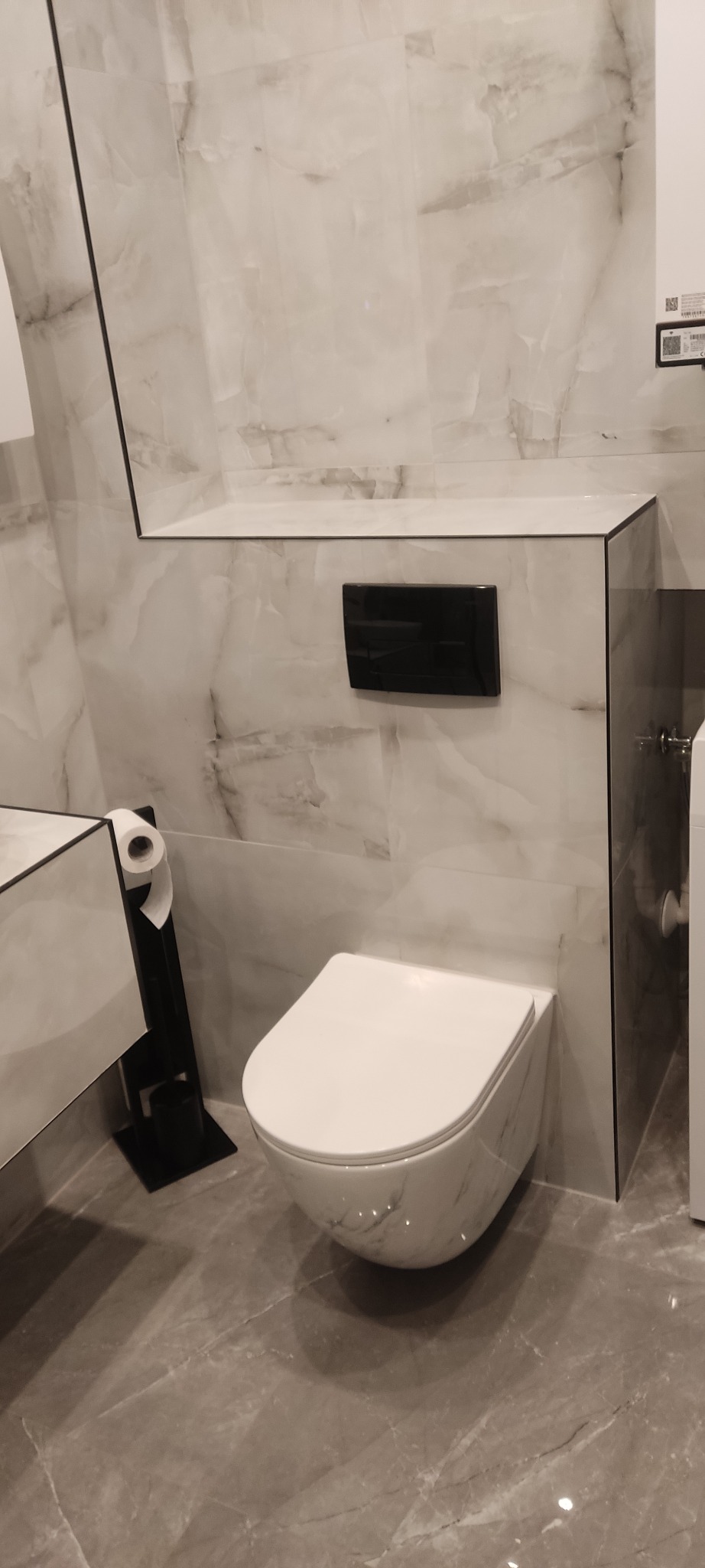 Nowoczesna toaleta podwieszana z czarnym przyciskiem spłukującym na ścianie wyłożonej marmurowymi płytkami, obok czarny stojak na papier toaletowy, szare marmurowe płytki podłogowe.