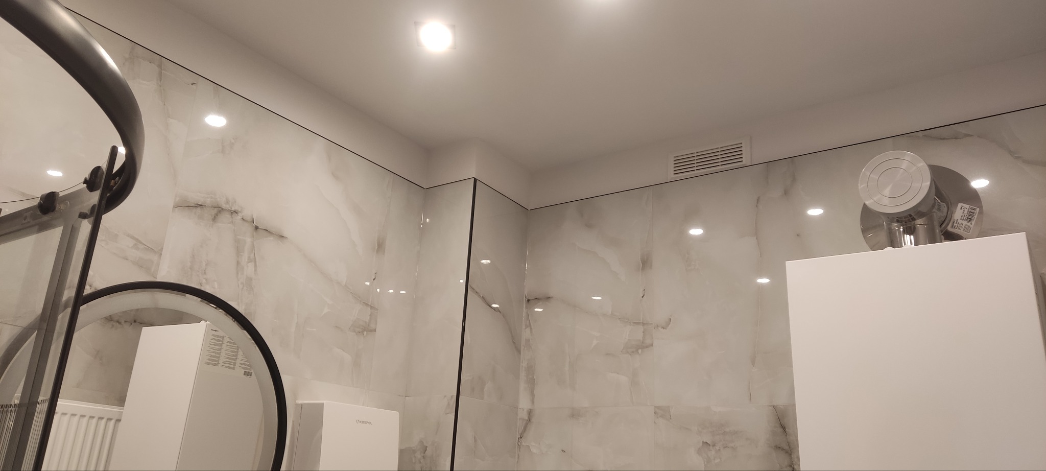 Wykończona łazienka z marmurowymi płytkami, kabiną prysznicową z czarnymi elementami i białymi szafkami, oświetlona punktowymi lampami sufitowymi.