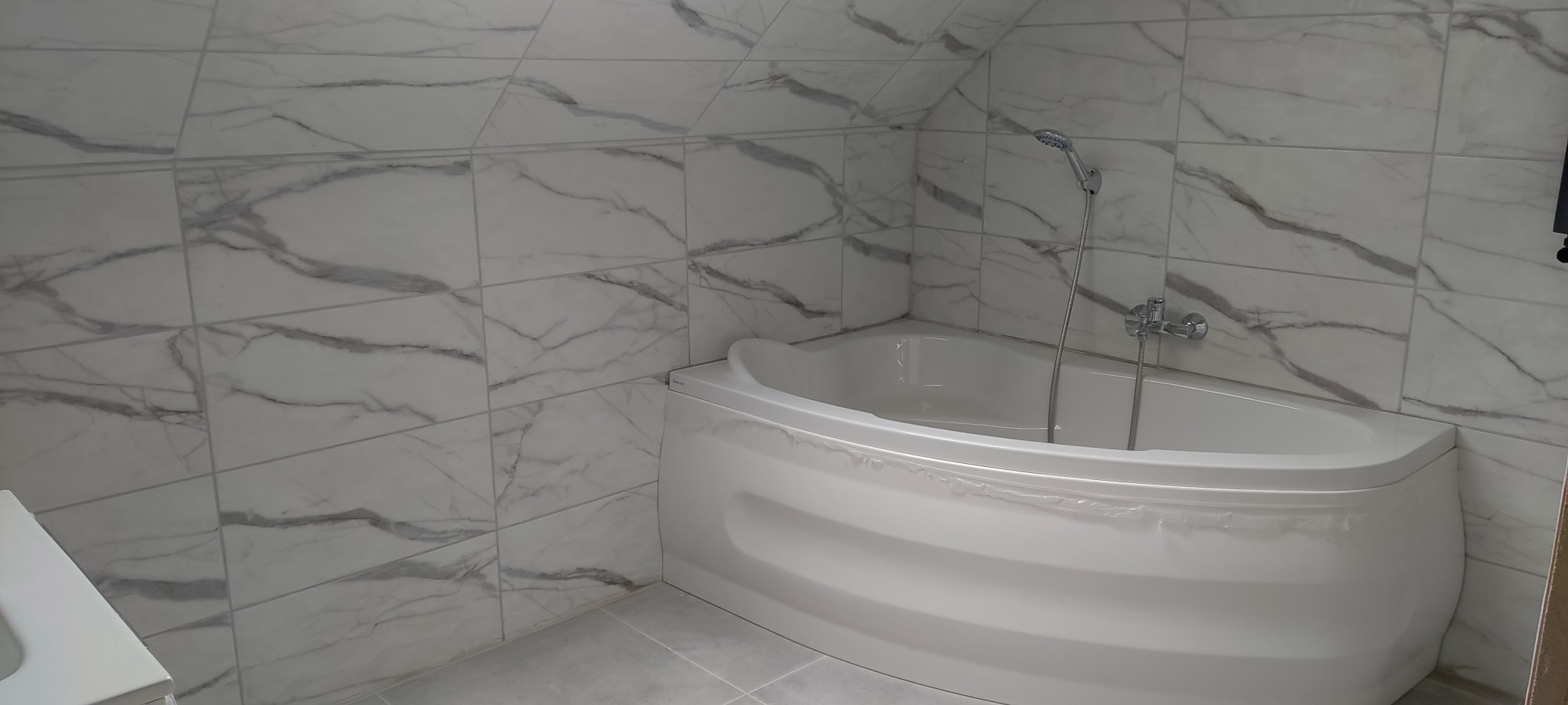 Wanna narożna z hydromasażem, obłożona białymi płytkami imitującymi marmur, zamontowana w łazience na poddaszu ze skośnym sufitem.