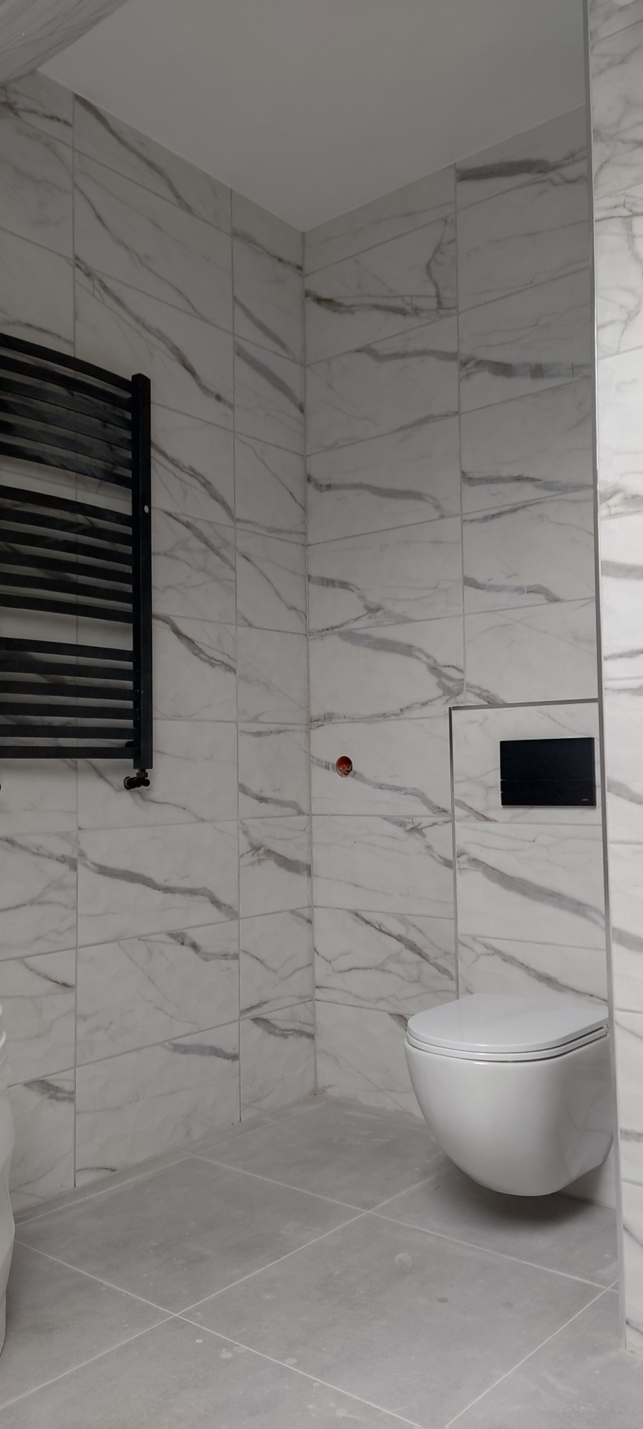 Wnętrze łazienki z płytkami imitującymi marmur na ścianach, wisząca toaleta z czarnym panelem sterującym, grzejnik drabinkowy w kolorze czarnym po lewej stronie, widoczne niedokończone prace...