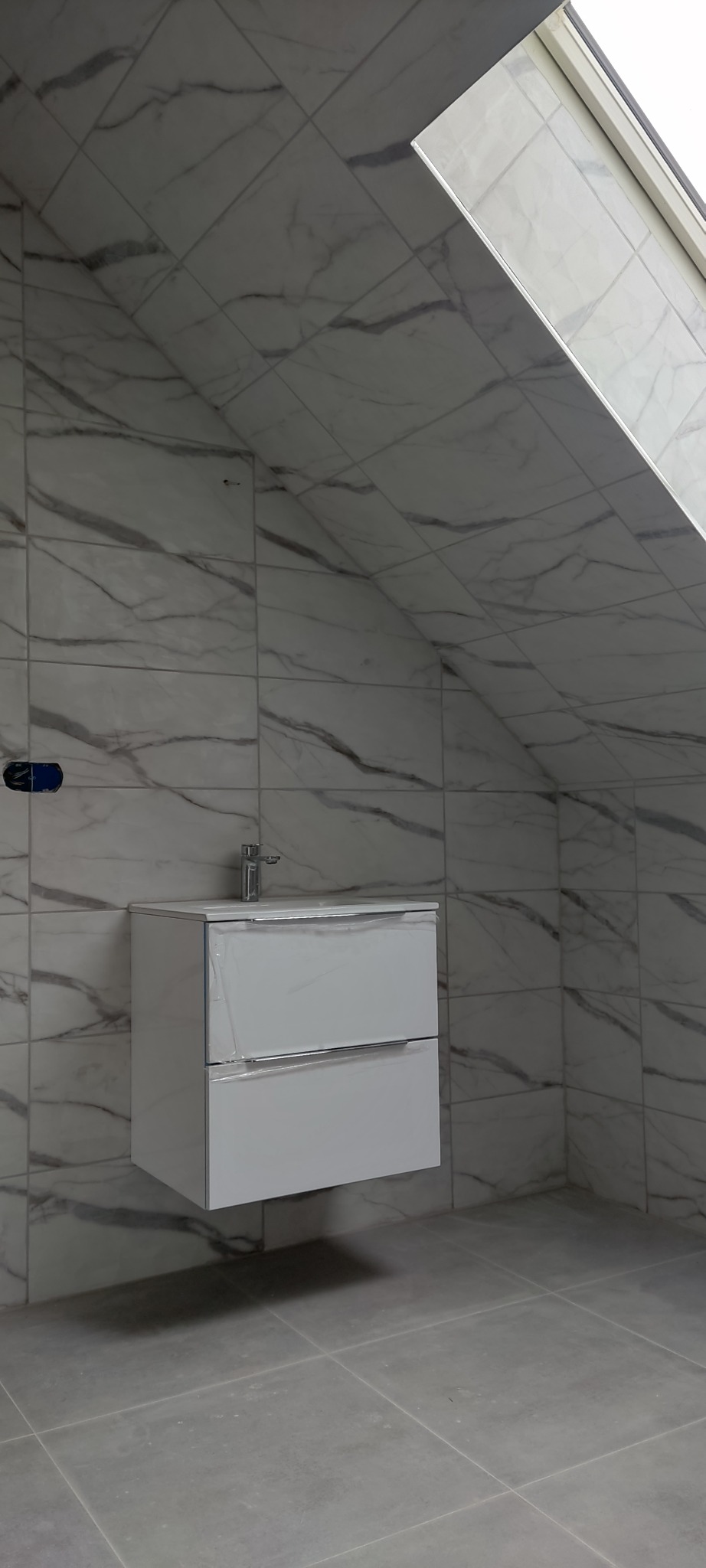 Wykończona łazienka na poddaszu z białą szafką pod umywalką i marmurowymi płytkami na ścianach i skośnym suficie z oknem dachowym.