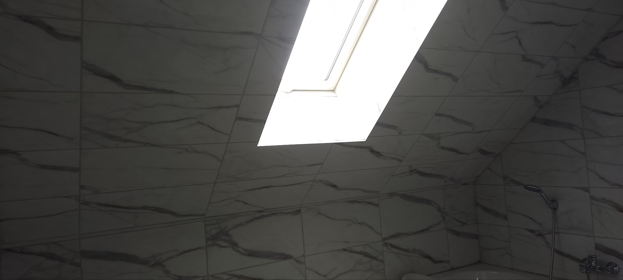 Wykończona łazienka na poddaszu z oknem dachowym, płytki imitujące marmur na ścianach, widoczna słuchawka prysznicowa z baterią.