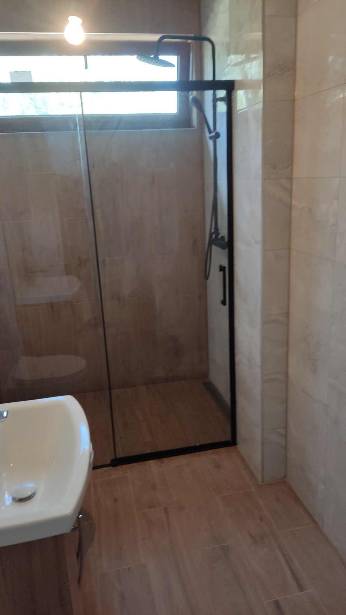 Nowoczesna kabina prysznicowa z czarną ramą i deszczownicą, widok na umywalkę i okno, jasne płytki imitujące drewno na ścianach i podłodze.