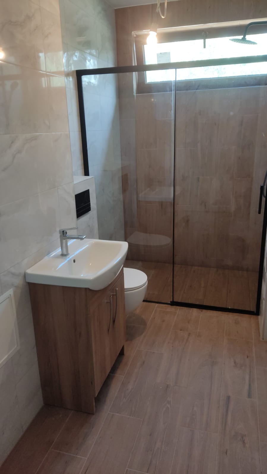 Nowoczesna łazienka z kabiną prysznicową typu walk-in z czarną ramą, umywalką na szafce z drewnopodobnym wykończeniem i toaletą podwieszaną, wykończona płytkami imitującymi drewno na podłodze...