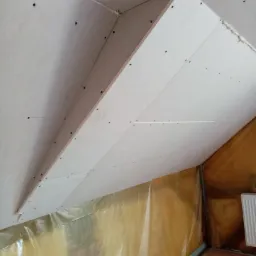 Skośny sufit na poddaszu w trakcie montażu płyt gipsowo-kartonowych, widoczne wkręty i szpachlowanie spoin, zabezpieczona folią podłoga.