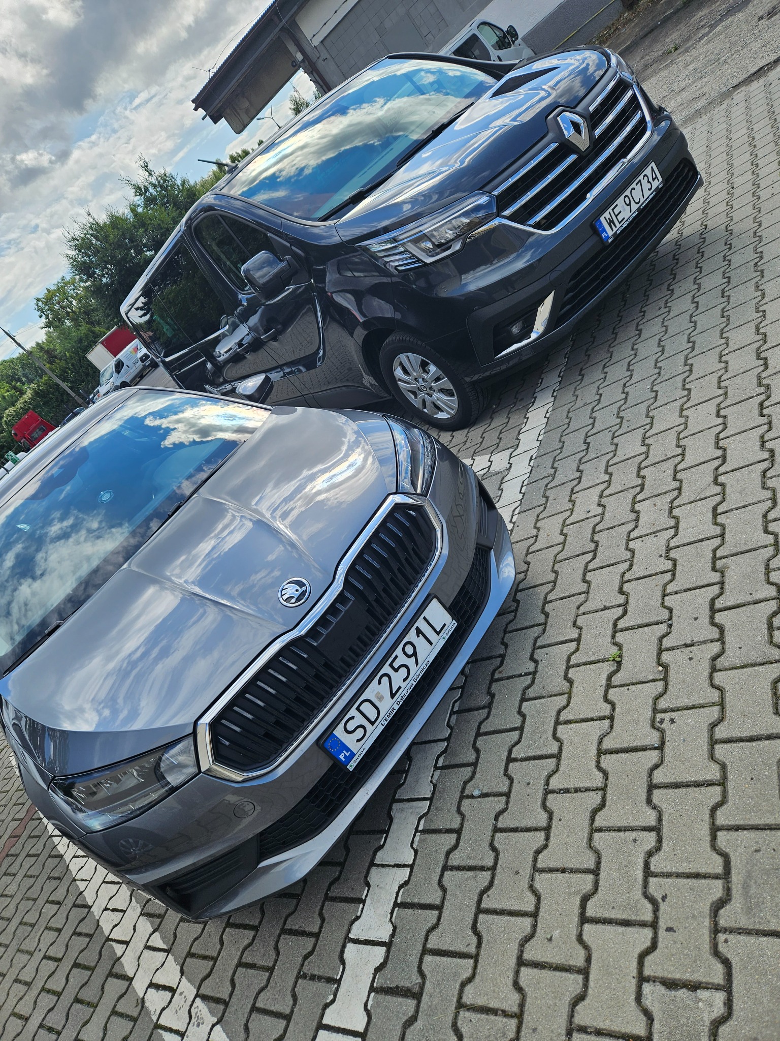 Dwa samochody: szary Skoda i ciemny Renault, zaparkowane na brukowanym placu, z odbiciem nieba w szybach.