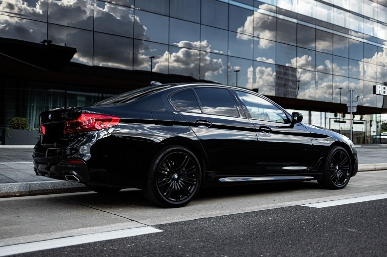 Czarny, luksusowy sedan marki BMW zaparkowany na tle nowoczesnego budynku ze szklaną fasadą odbijającą chmury.