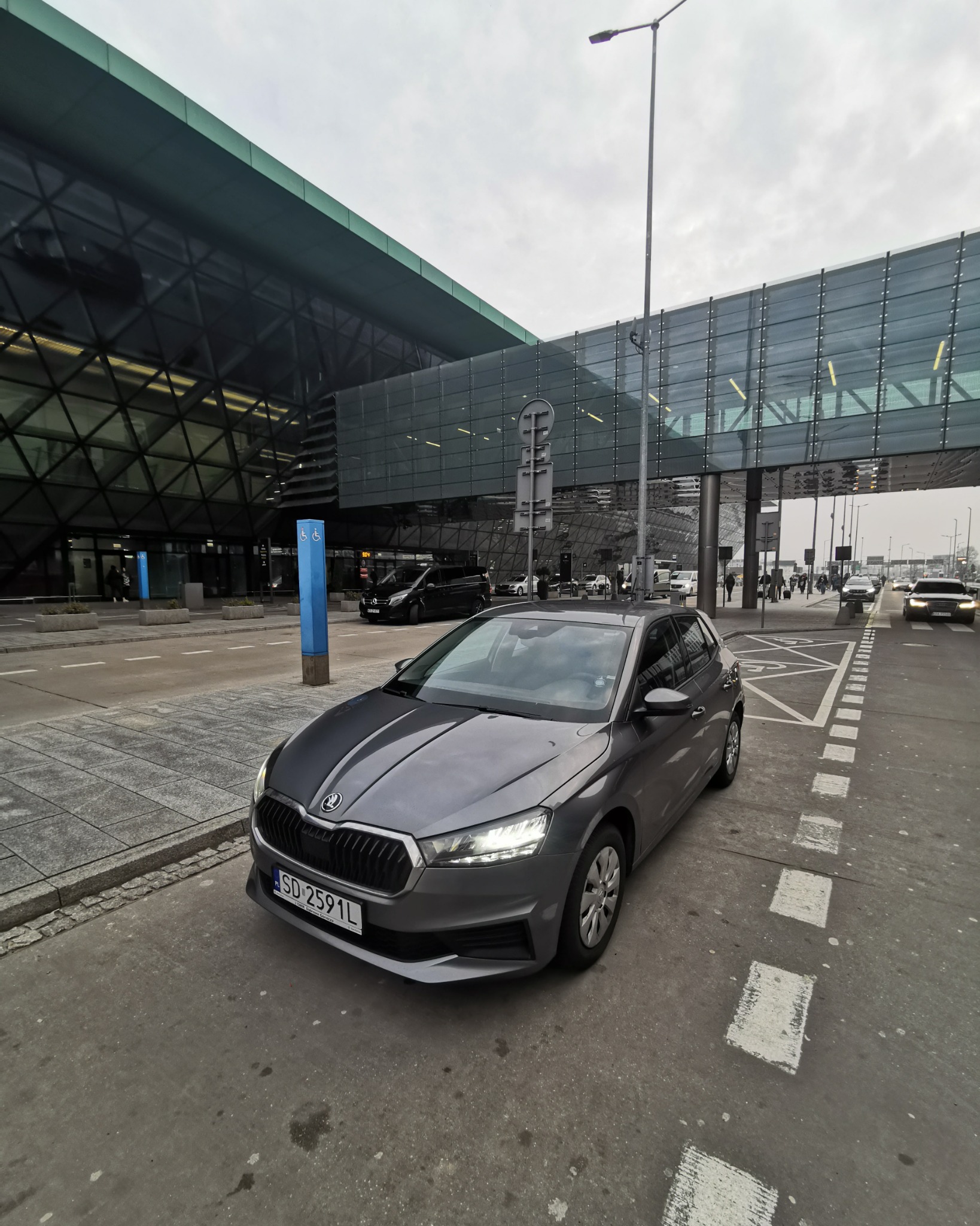 Szara Skoda Fabia zaparkowana przed nowoczesnym budynkiem lotniska z przeszklonymi fasadami i nadziemnym przejściem dla pieszych.