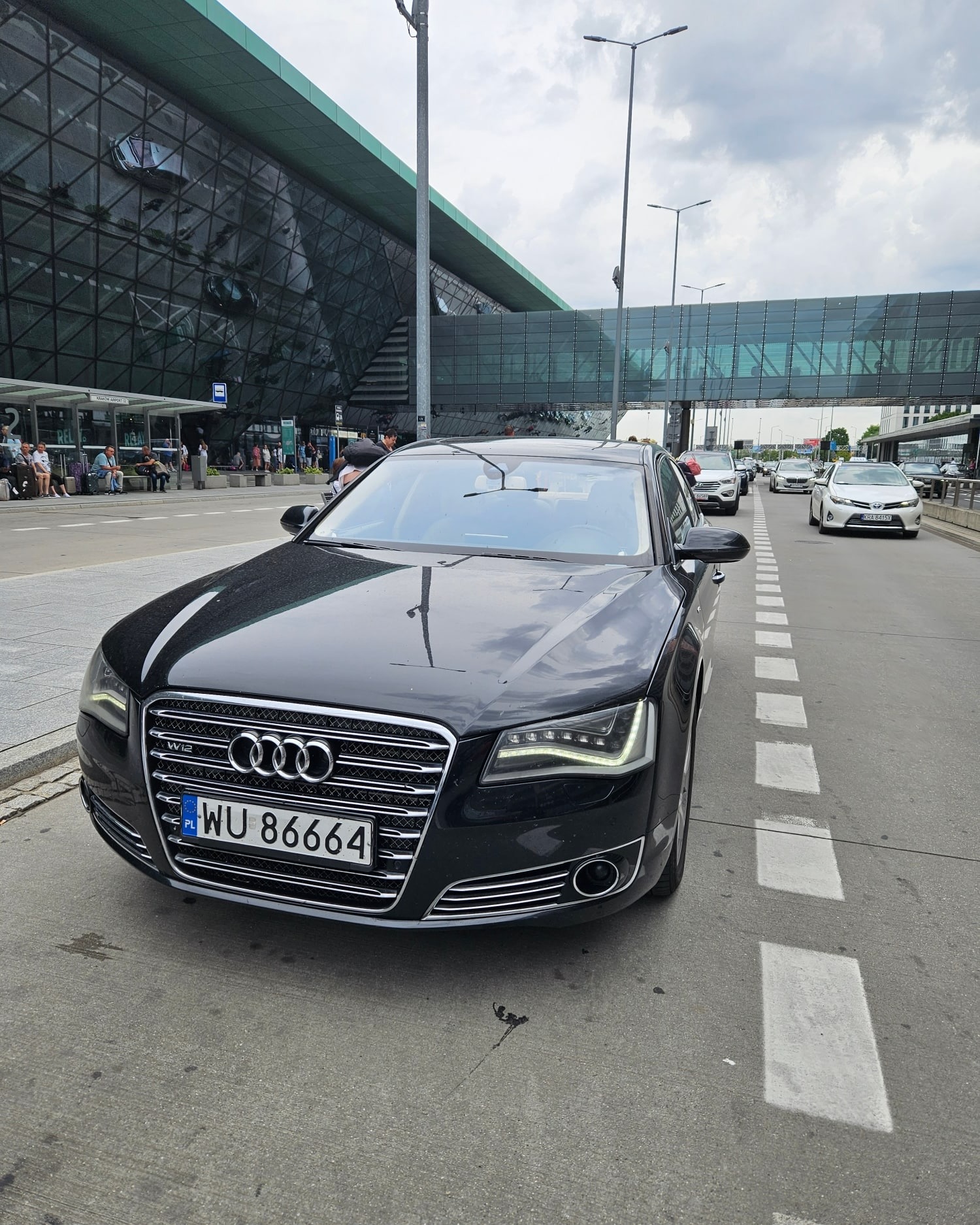 Czarna limuzyna Audi z widoczną tablicą rejestracyjną WU 86664 na tle nowoczesnego budynku lotniska z przeszkloną fasadą i pasażami dla pieszych.