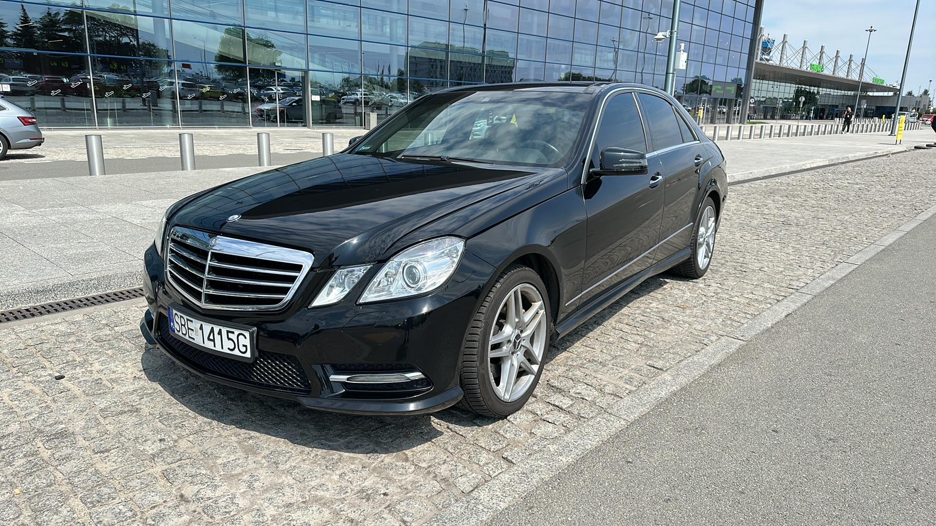 Czarny Mercedes klasy E zaparkowany na kostce brukowej przed nowoczesnym budynkiem z przeszkloną fasadą, prawdopodobnie lotniskiem, w słoneczny dzień.