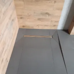 Nowoczesny odpływ liniowy w szarej, kwadratowej posadzce z płytek, otoczony ścianami wyłożonymi płytkami imitującymi drewno i jasnoszarą płytką.