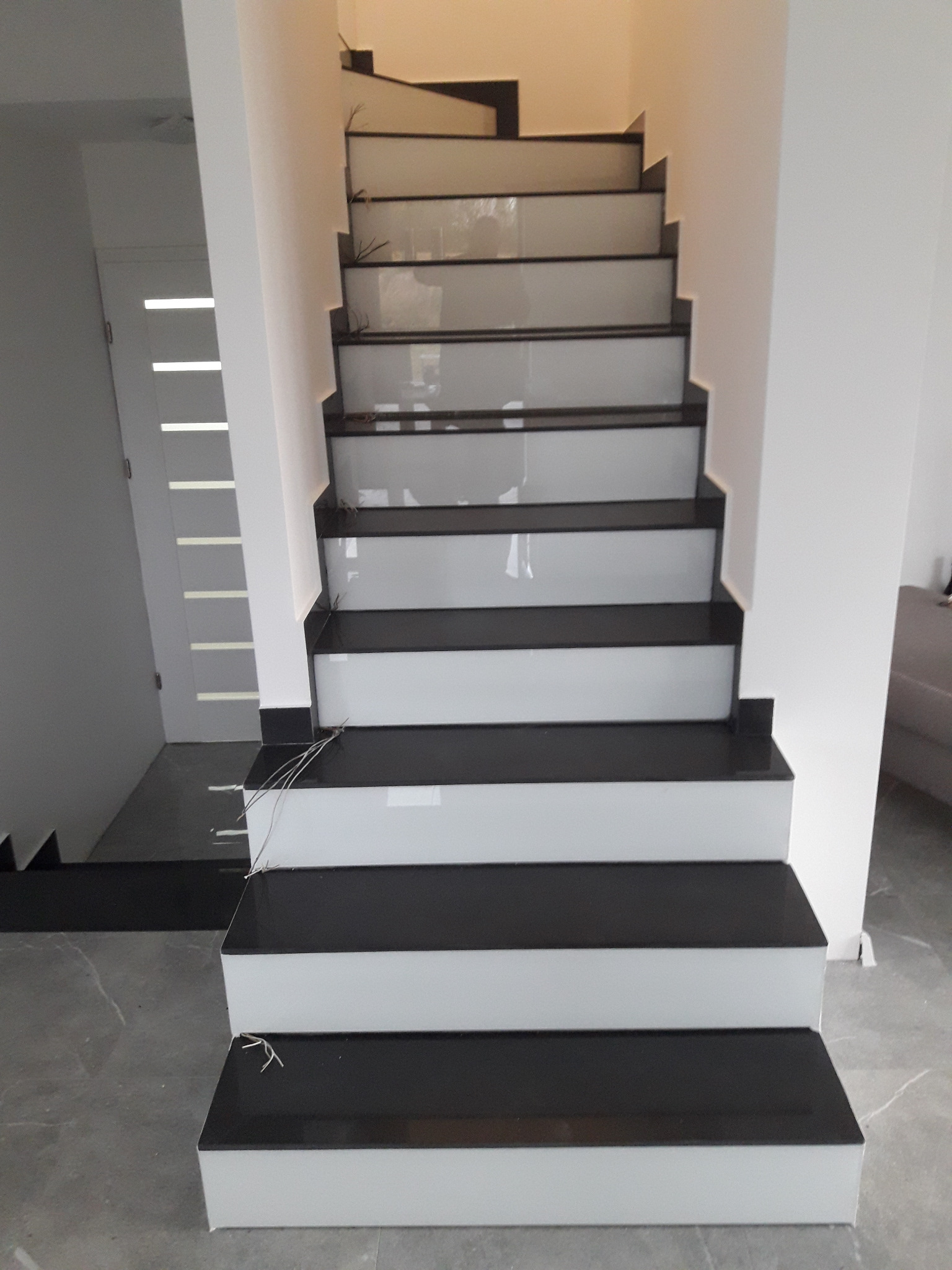 Nowoczesne schody wewnętrzne z czarnymi stopniami i białymi podstopnicami, widoczne kable instalacyjne przy dolnych stopniach, szara marmurowa podłoga, białe ściany i drzwi w tle.