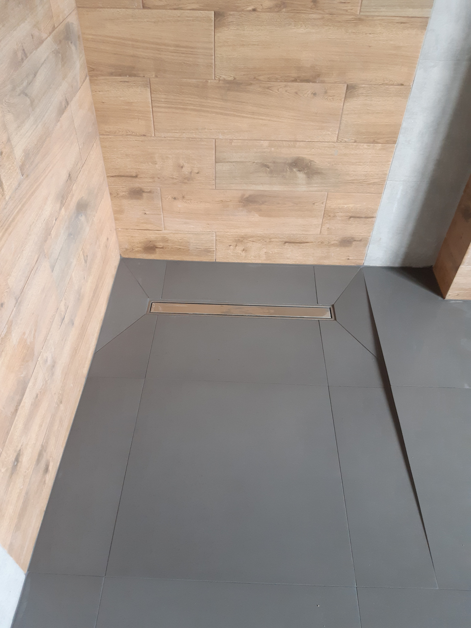 Nowoczesny odpływ liniowy w szarej, kwadratowej posadzce z płytek, otoczony ścianami wyłożonymi płytkami imitującymi drewno i jasnoszarą płytką.