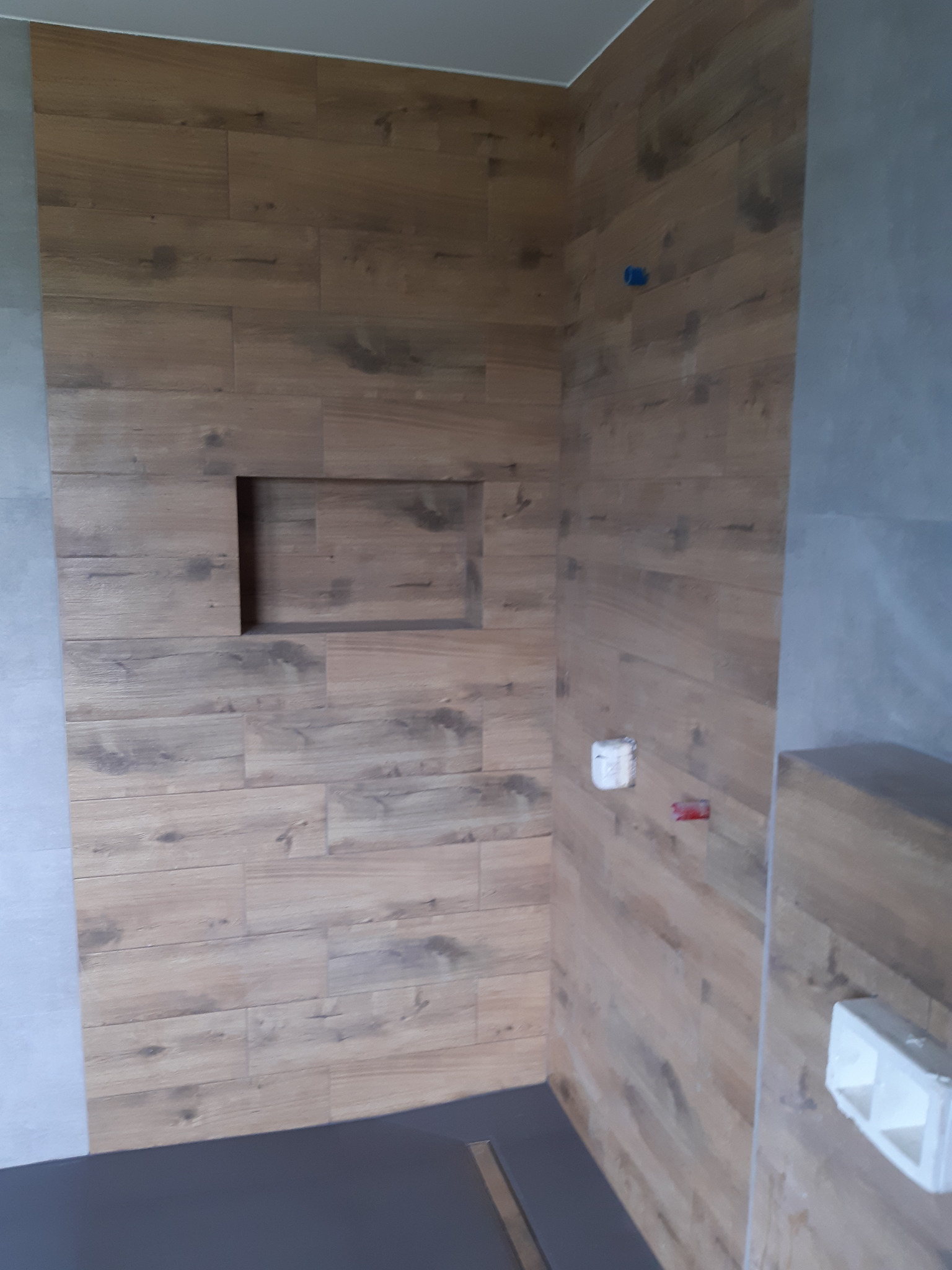 Wykończona kabina prysznicowa z drewnopodobnymi płytkami, wnęką w ścianie i szarą posadzką z odpływem liniowym.