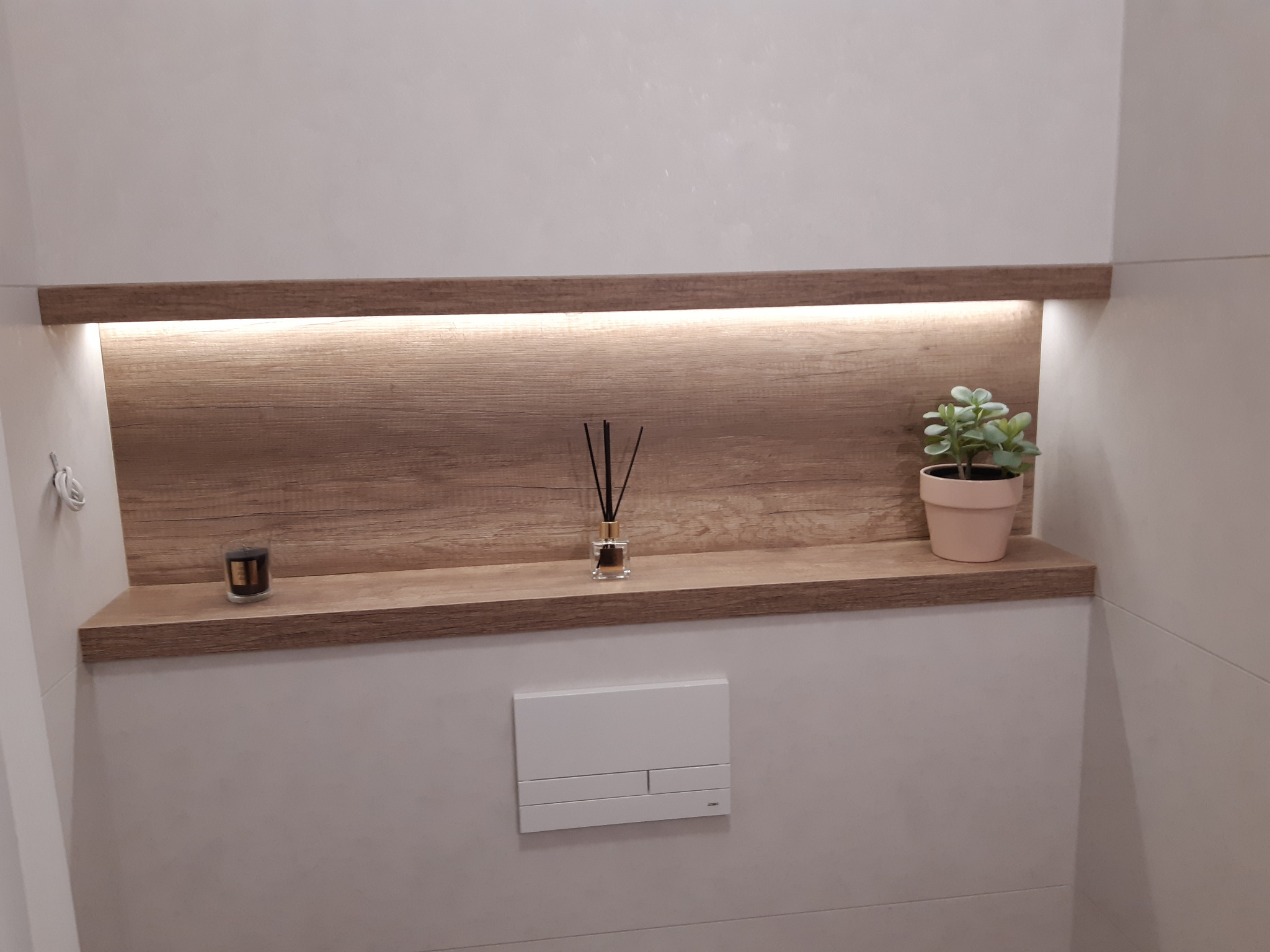 Nowoczesna toaleta z wbudowaną półką z podświetleniem LED, na której stoją świeczka zapachowa, dyfuzor zapachowy i mała roślina doniczkowa.