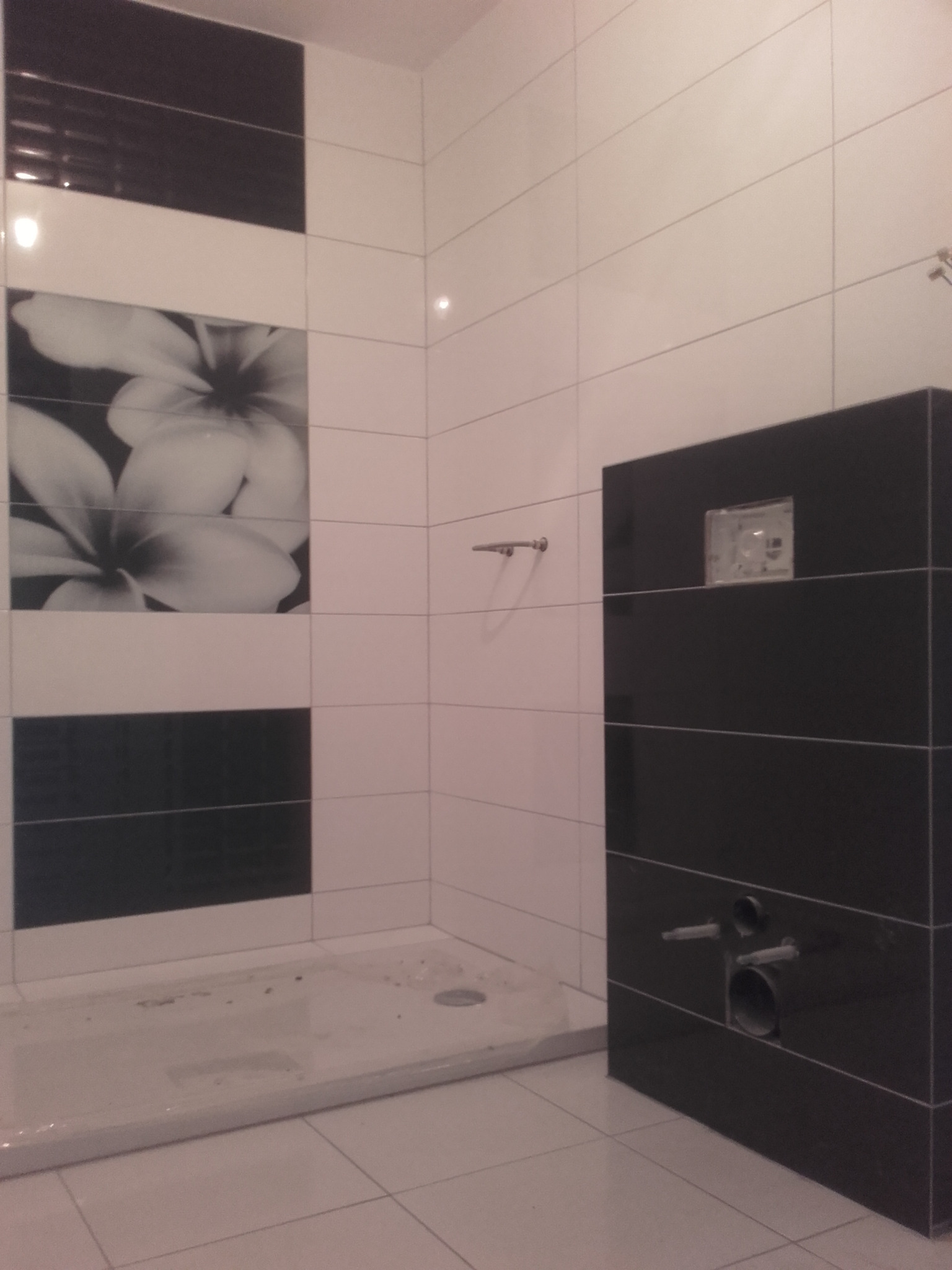 Nowoczesna łazienka w trakcie wykańczania, z białymi płytkami i dekoracyjnym panelem z motywem kwiatowym, widoczna zabudowa stelaża WC z czarnych płytek, surowy brodzik.