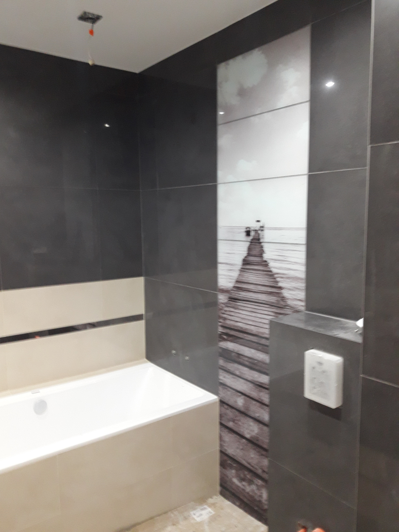 Wykończona łazienka z wanną, ciemnymi płytkami i dekoracyjnym panelem ściennym z motywem molo na tle morza, widoczny biały włącznik światła.