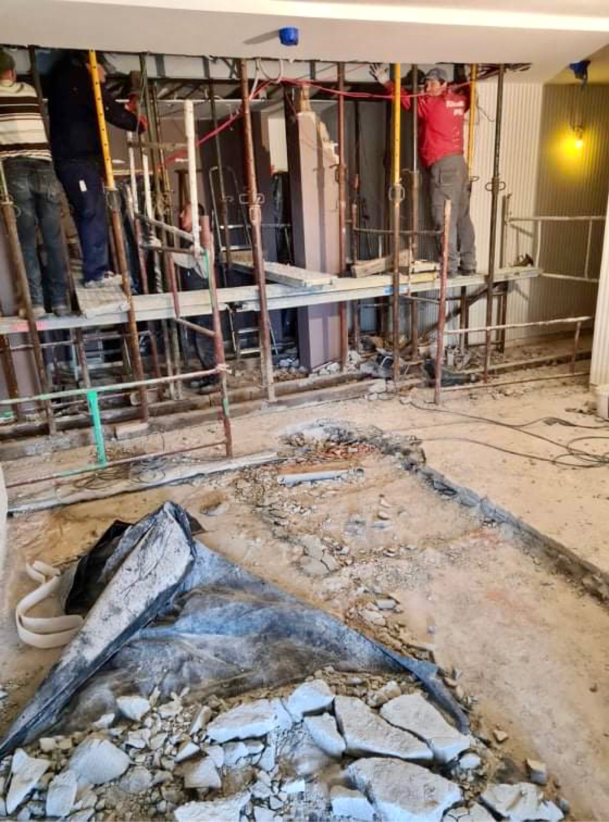 Wnętrze w trakcie remontu: pracownicy na rusztowaniach, odsłonięte instalacje pod sufitem, gruz i zabezpieczenia na podłodze.