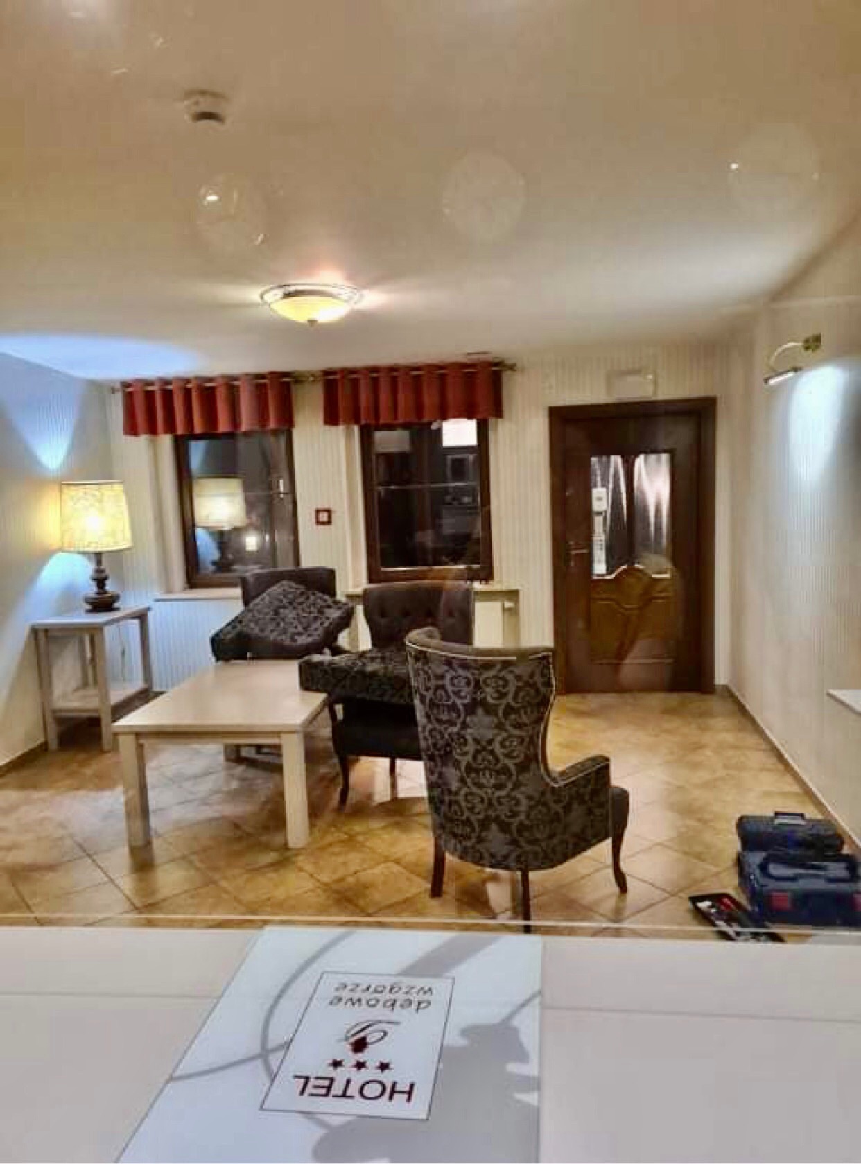 Wnętrze hotelu z fotelami w stylu vintage, stolikiem i narzędziami w rogu, widoczne przez szybę z logo hotelu.