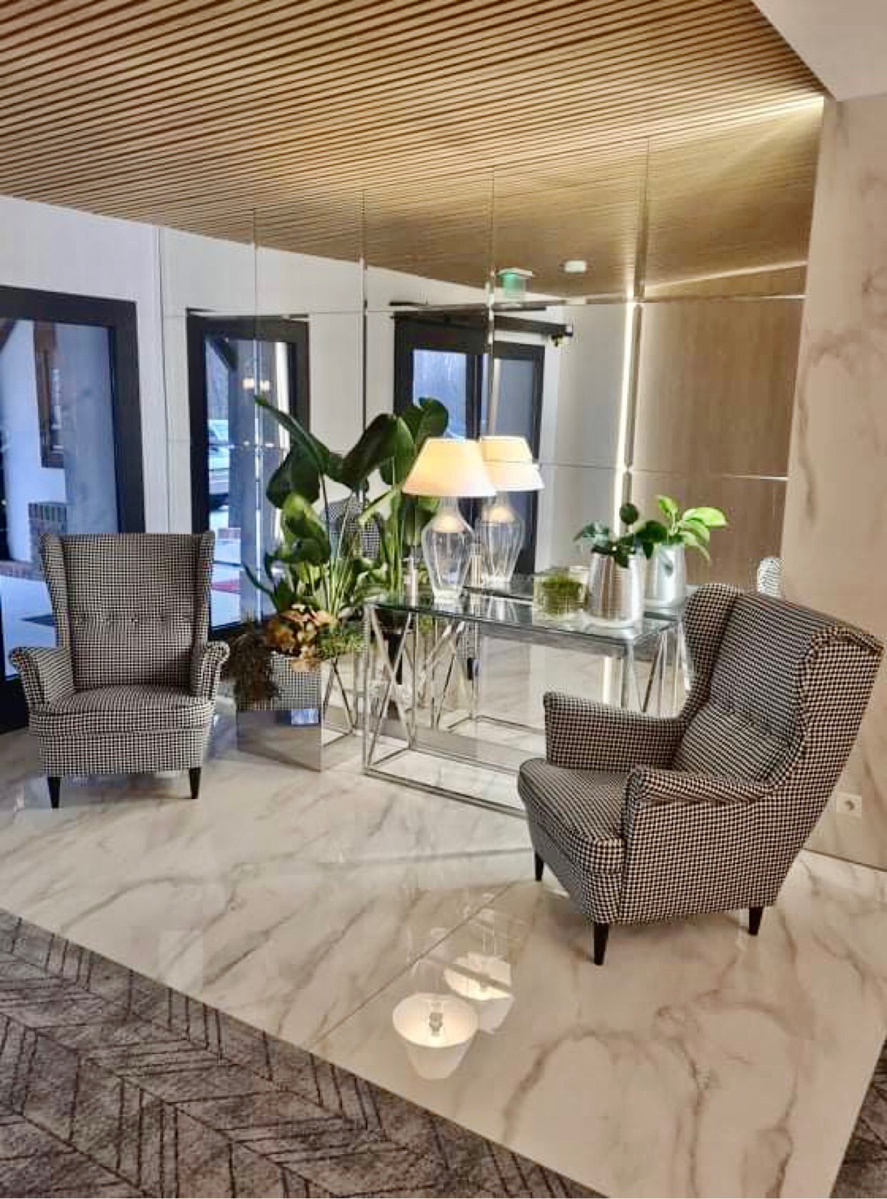 Eleganckie wnętrze holu z marmurową podłogą, lustrzaną ścianą, dwoma fotelami w pepitkę i stolikiem ze szklanym blatem oraz roślinami i lampami.