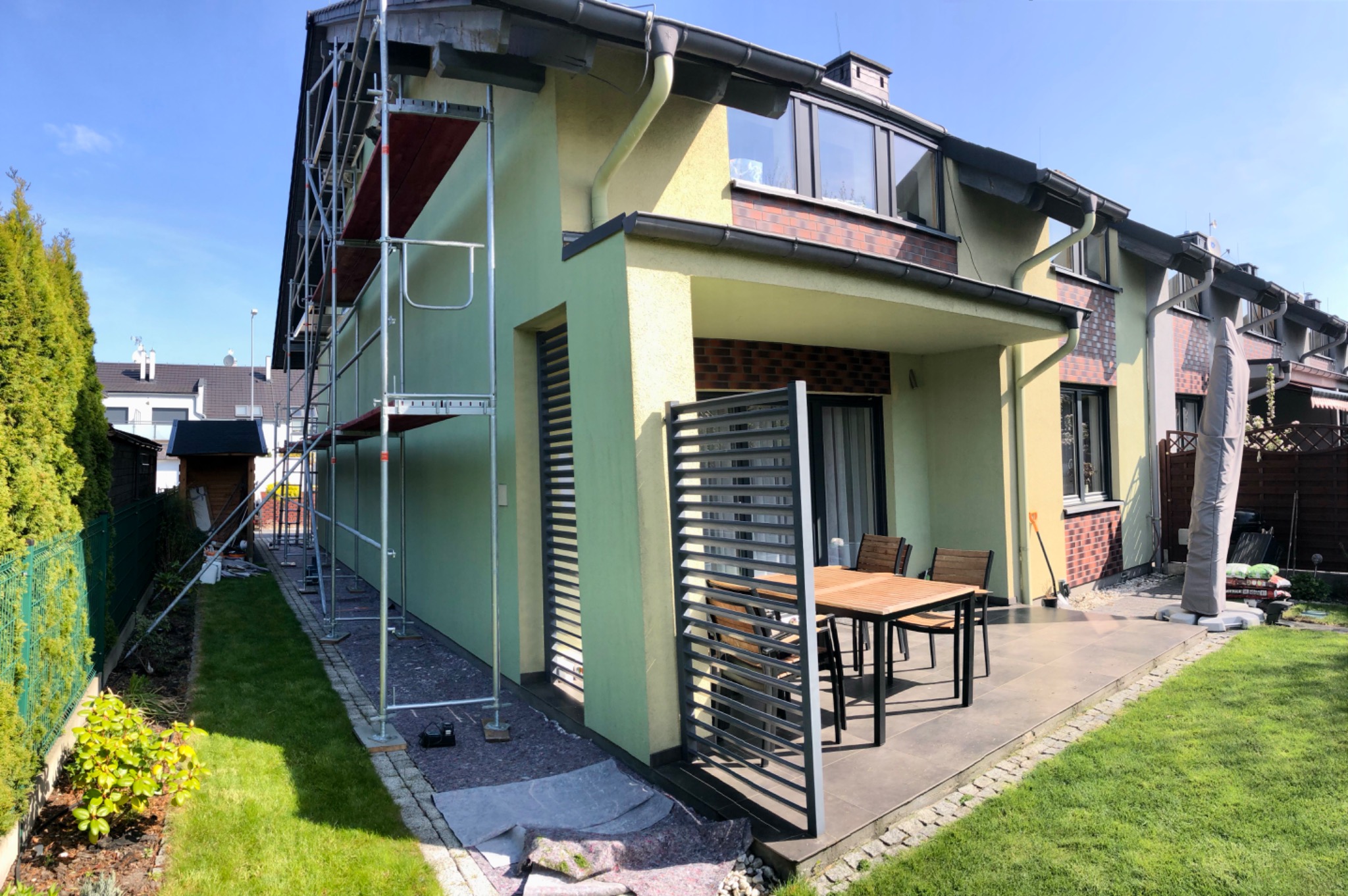 Dom jednorodzinny w trakcie prac elewacyjnych, widoczne rusztowania przy ścianie, zabezpieczona folią przestrzeń wokół domu, świeżo pomalowana elewacja w odcieniach zieleni i beżu.