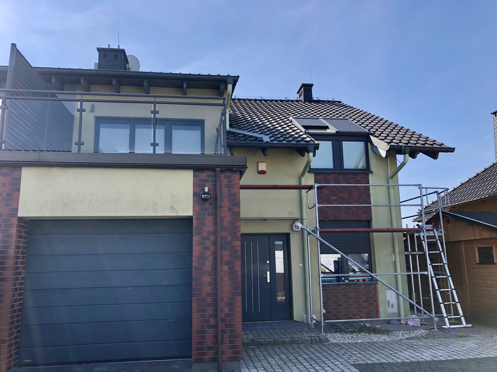 Dwukondygnacyjny dom jednorodzinny z balkonem i garażem, częściowo otoczony rusztowaniem, z fragmentami elewacji wykończonymi cegłą klinkierową.