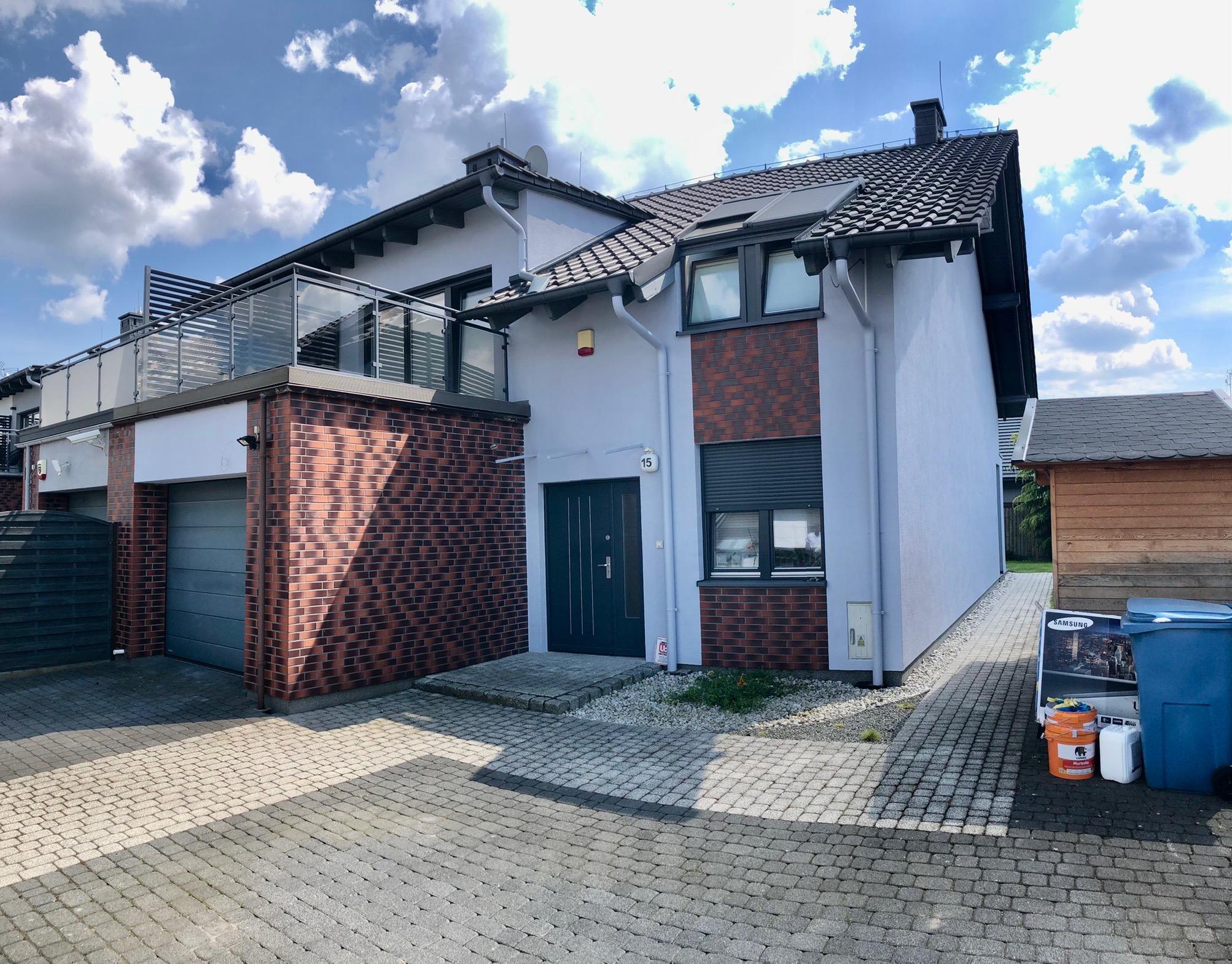 Dwukondygnacyjny dom z balkonem, szarą elewacją i elementami z czerwonej cegły, widok z zewnątrz na podjeździe z kostki brukowej, z boku widoczny pojemnik na śmieci i karton po telewizorze.