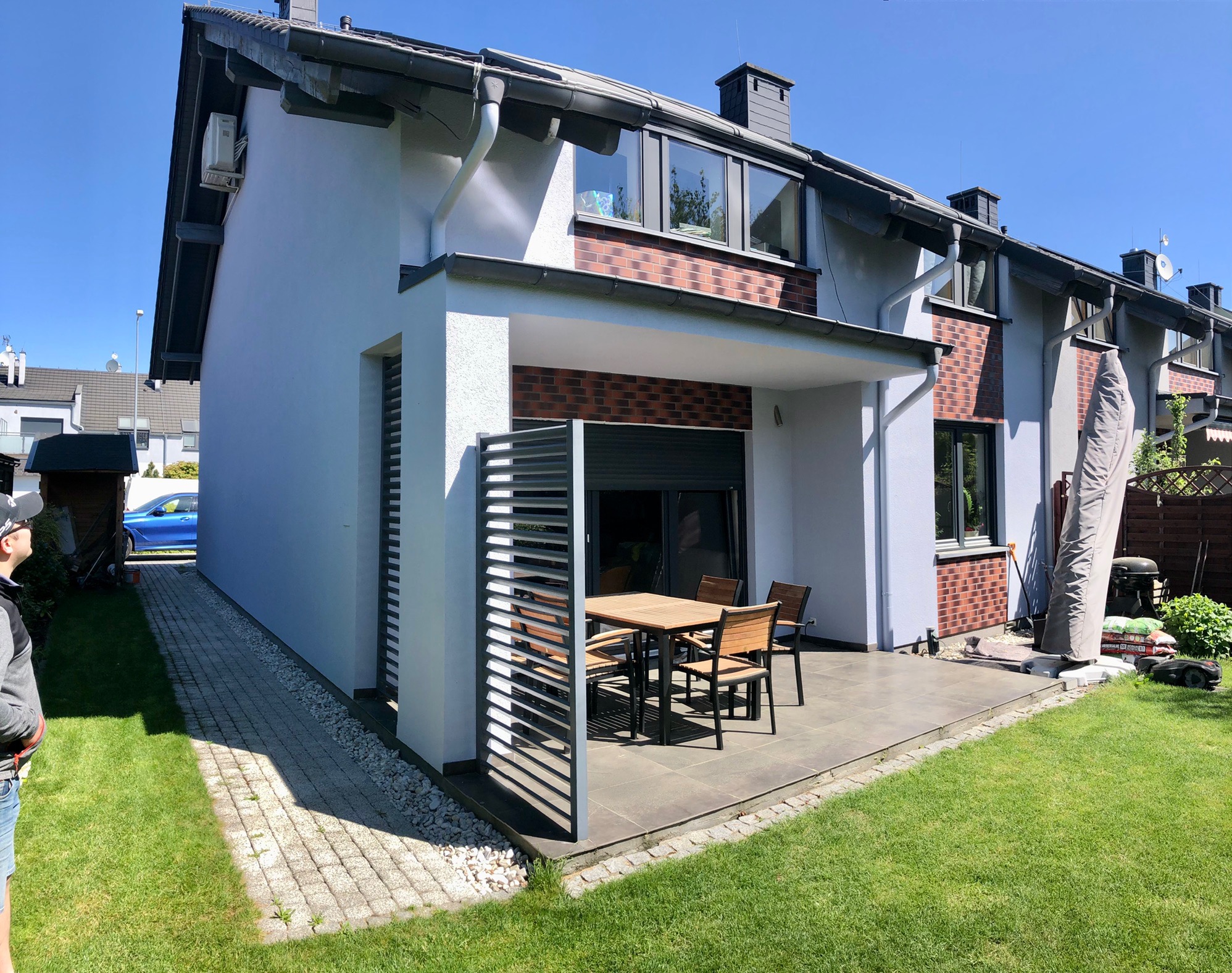 Otynkowana elewacja domu szeregowego z ceglaną dekoracją, widoczny taras z meblami ogrodowymi i zadbany trawnik.