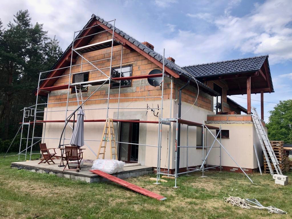 Dwukondygnacyjny dom w trakcie prac elewacyjnych, widoczne rusztowania, częściowo otynkowana fasada, odkryta cegła na górnej kondygnacji, teren posesji z meblami ogrodowymi i narzędziami.