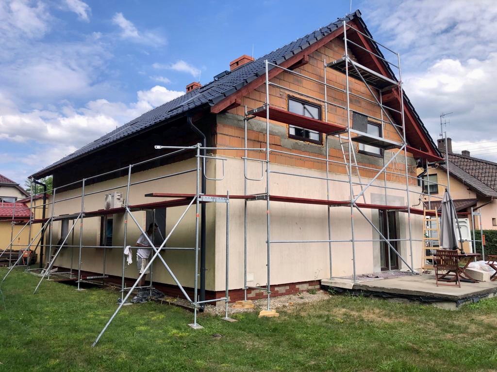 Dom jednorodzinny w trakcie prac elewacyjnych, widoczne rusztowania, fragmenty ocieplenia i tynku, ceglana ściana szczytowa, pochmurne niebo w tle.