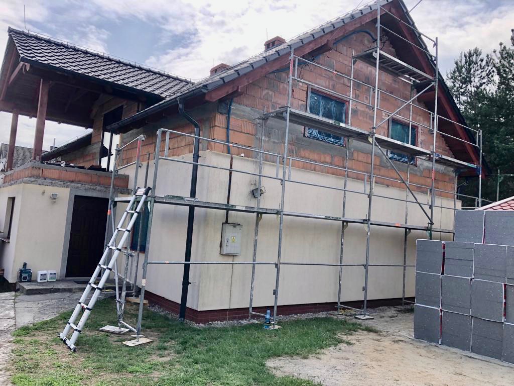 Dom w trakcie budowy, z częściowo otynkowaną elewacją, rusztowaniem i stosem płyt izolacyjnych obok.