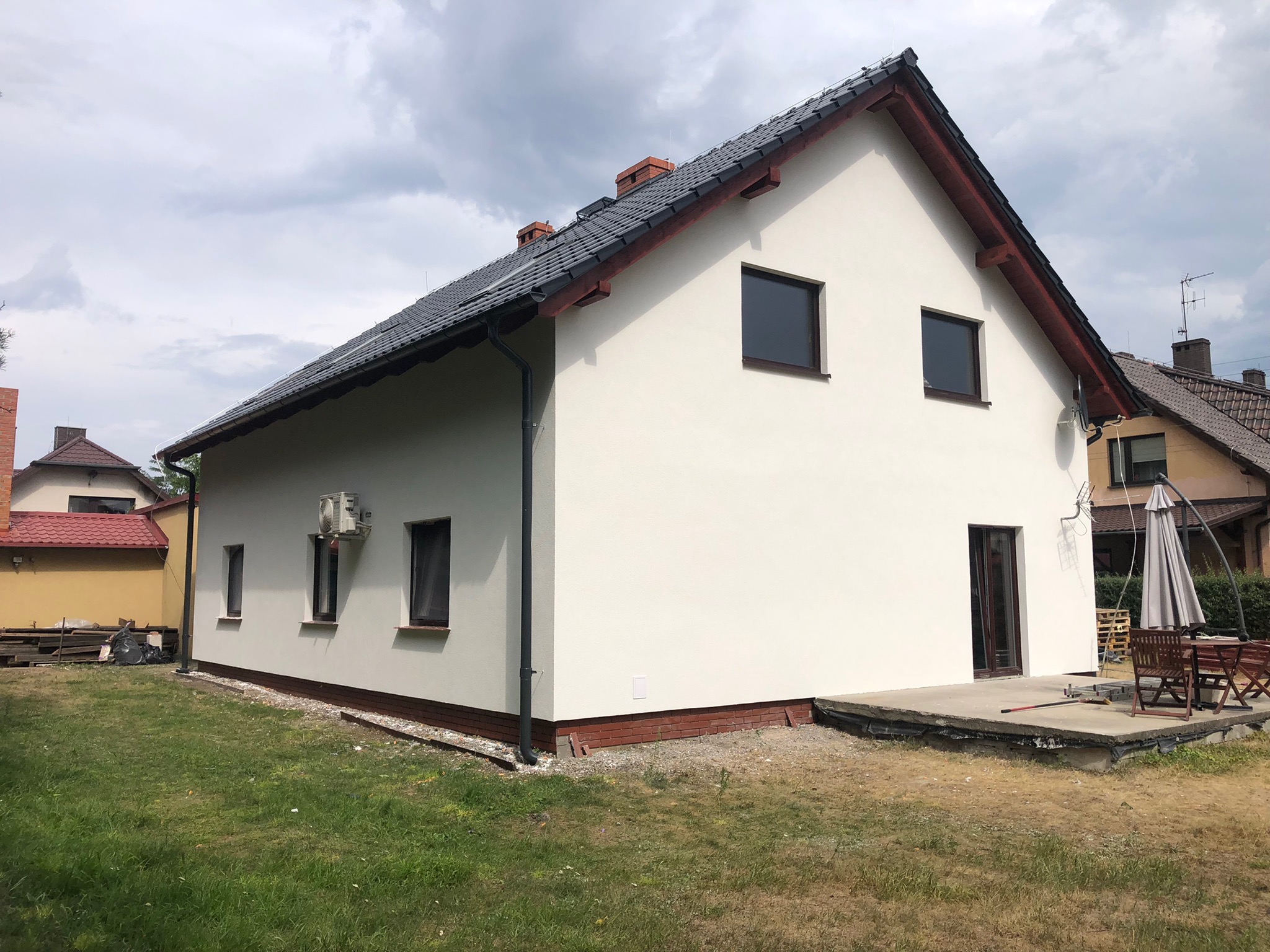 Świeżo otynkowany, biały dom jednorodzinny z ciemną dachówką, widok z boku na ogród w pochmurny dzień.