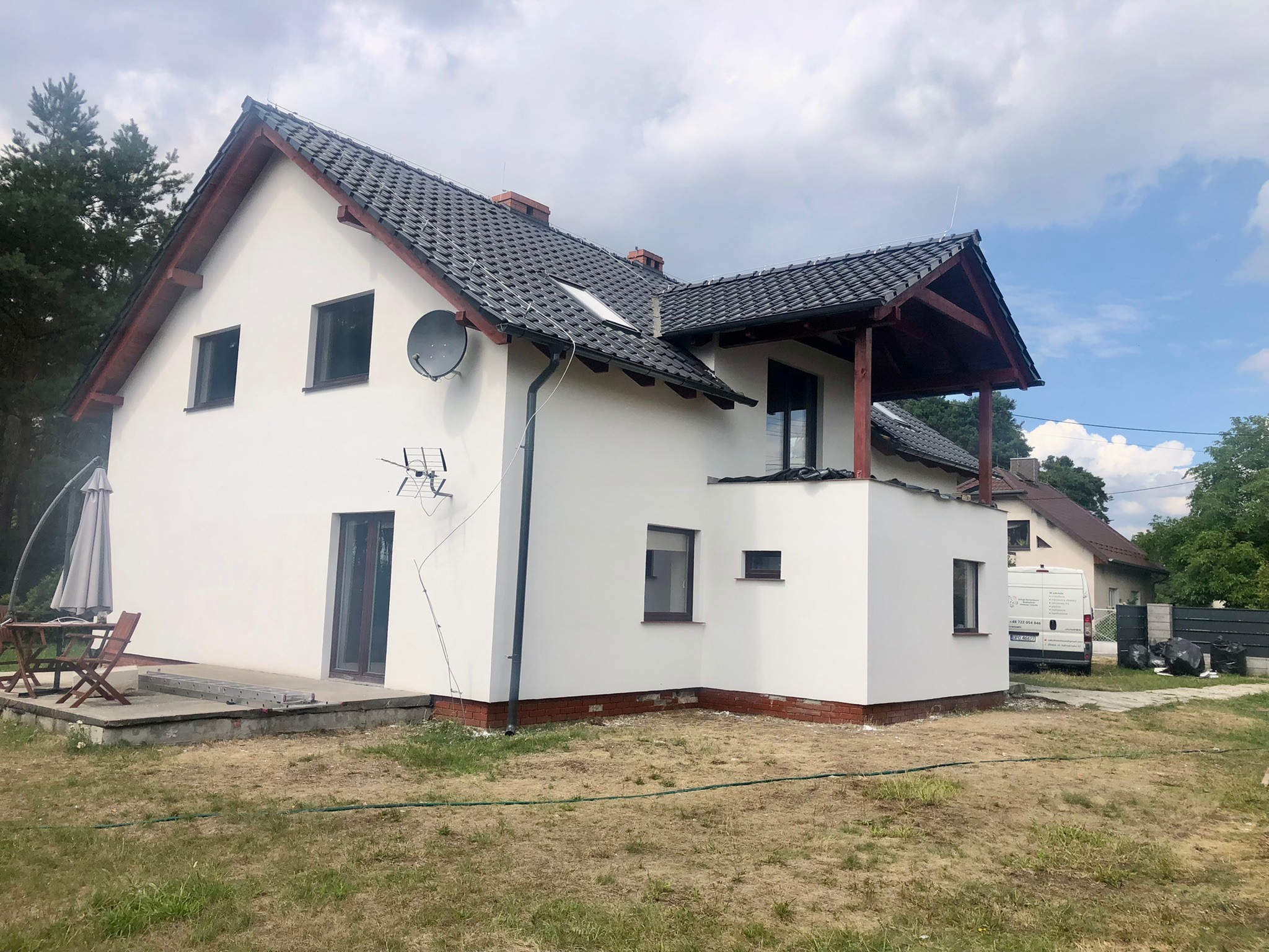 Biały dom jednorodzinny z szarą dachówką, widoczna antena satelitarna, okna z brązowymi ramami, częściowo ocieplona elewacja, na trawniku leży drabina, w tle zaparkowany biały bus.