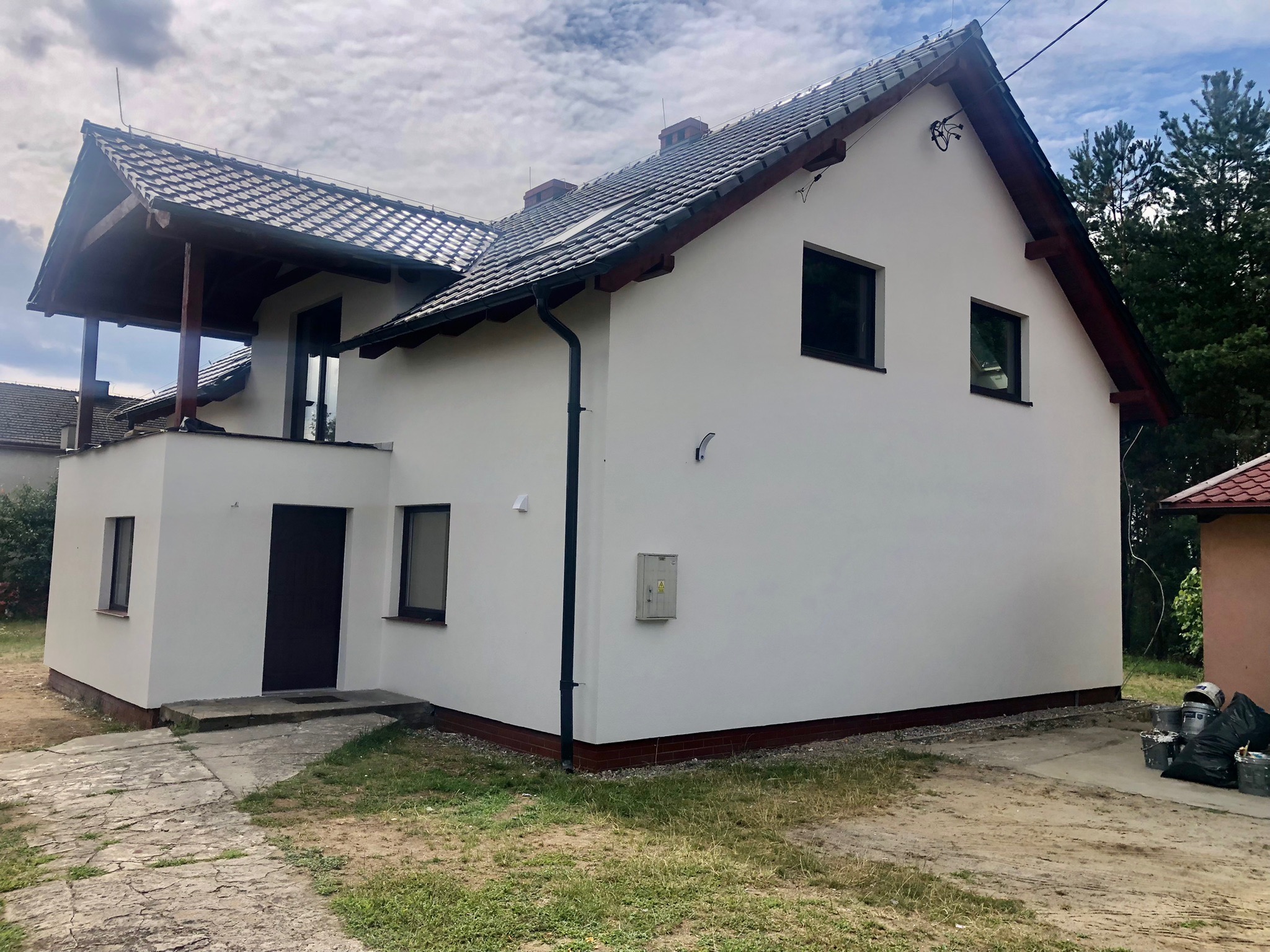 Dwupiętrowy dom z nową, białą elewacją, ciemną stolarką okienną i brązowym wykończeniem dachu, widoczny z perspektywy trawnika i ścieżki z kostki.