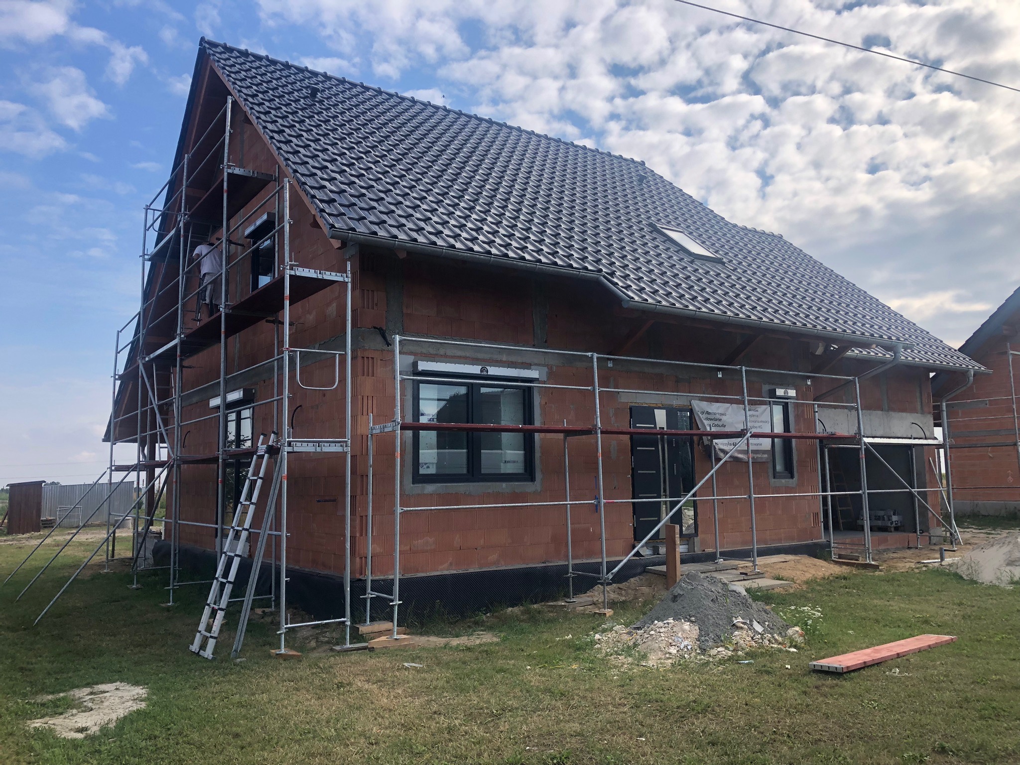 Dom w stanie surowym otwartym z rusztowaniem, widoczne okna i drzwi, dach pokryty dachówką, na trawniku leżą materiały budowlane.