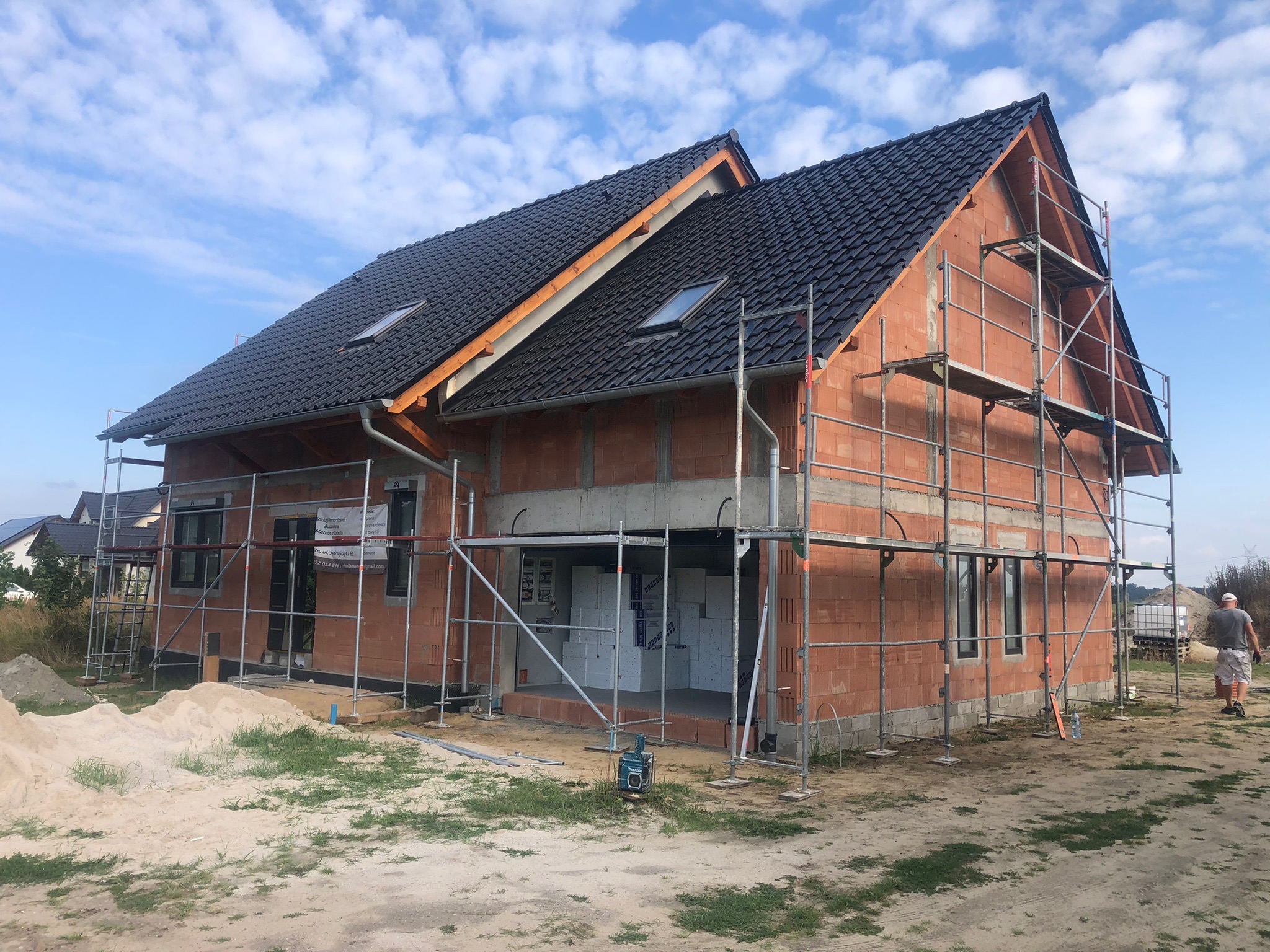 Niedokończony dom jednorodzinny w trakcie budowy, z widocznymi rusztowaniami, nowym dachem z dachówki ceramicznej i oknami, ściany z czerwonej cegły porotherm, na działce piasek i trawa.